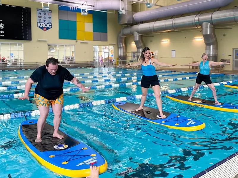 people standing up on a paddle board performing a warrior 2 pose during an AQUA STAND UP® class in a swimming pool