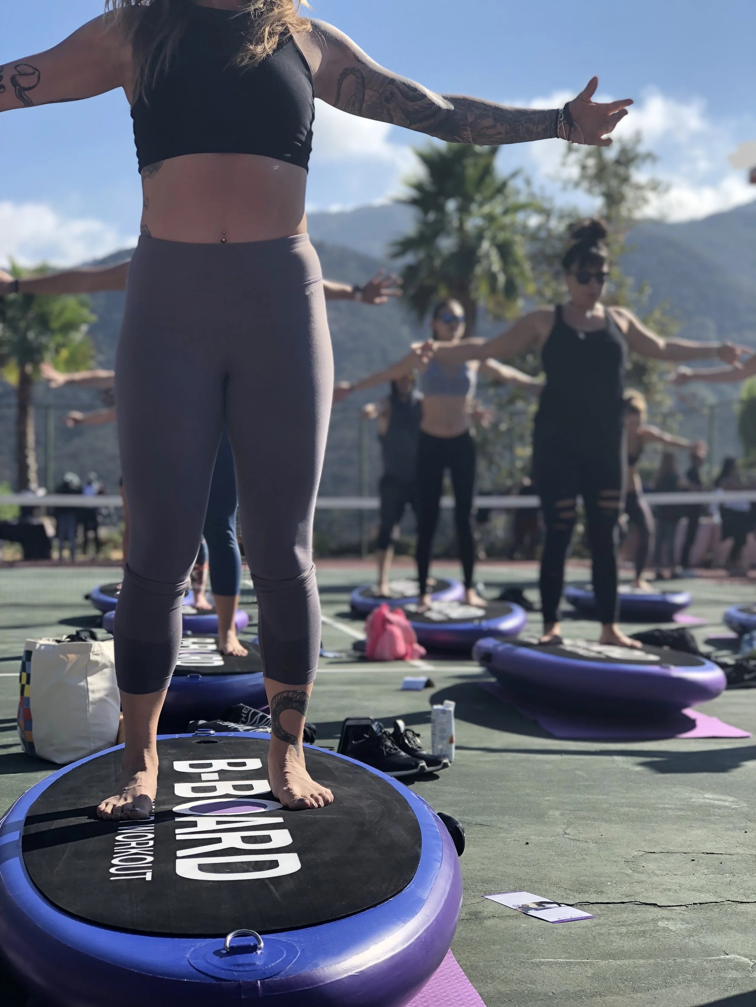 women practicing balance exercise on a balance board called B-BOARD® on a tennis court with other participants in background, blue sky and palm trees