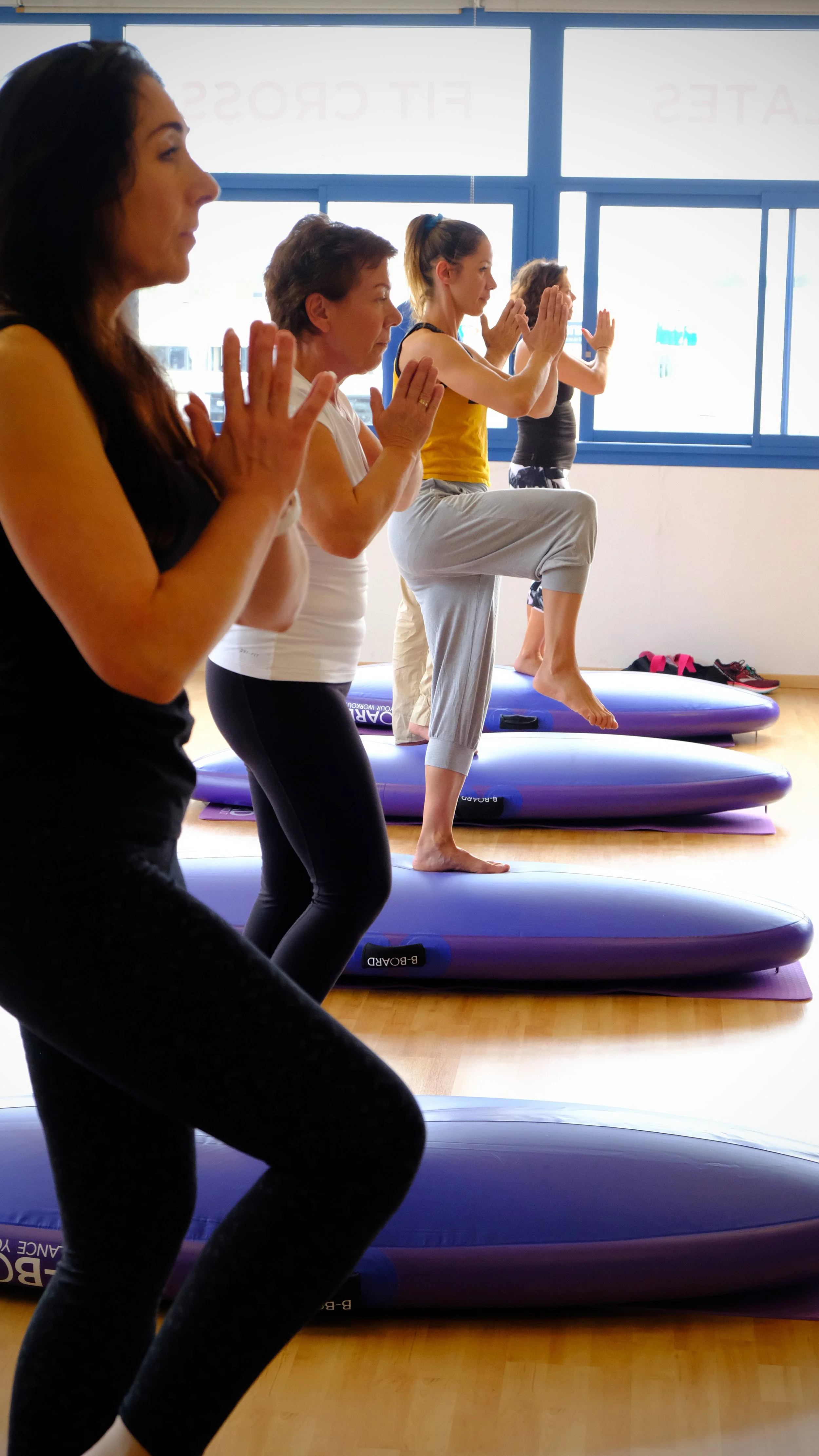 group of women performing a balance pose on the round part of the B-BOARD® with hands in prayer and one knee up