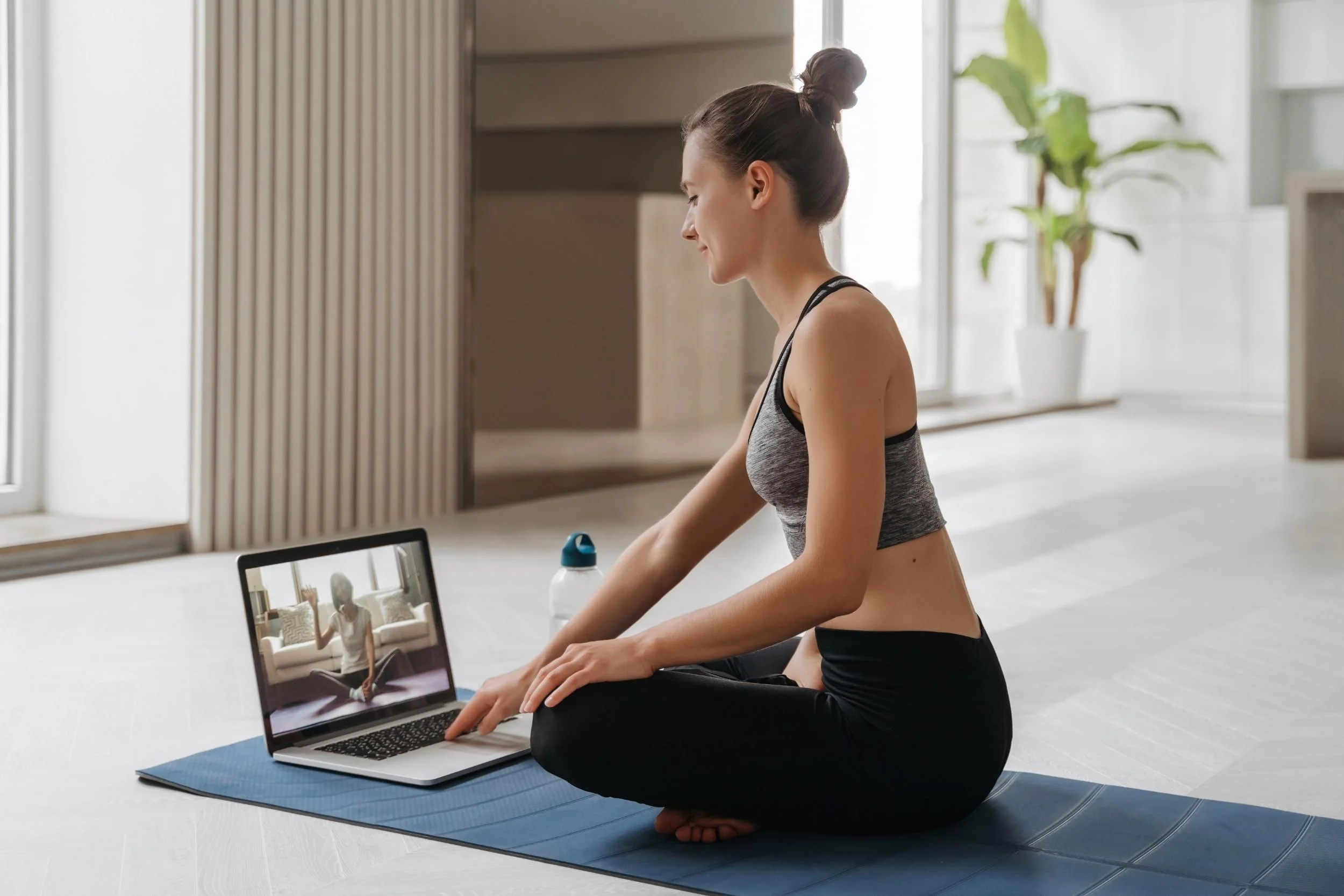 A woman in workout clothes sitting on a yoga mat, following an online yoga session on her laptop in a bright, modern room with large windows and a potted plant.