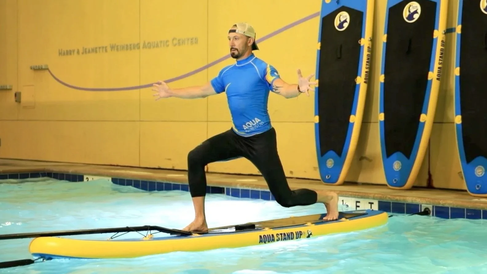 aquastandup instructor wearing a black leggins and a blue rashguard branded aquastandup performing a lunge on a paddle board in a indoor swimming pool with aqua stand up boards in background