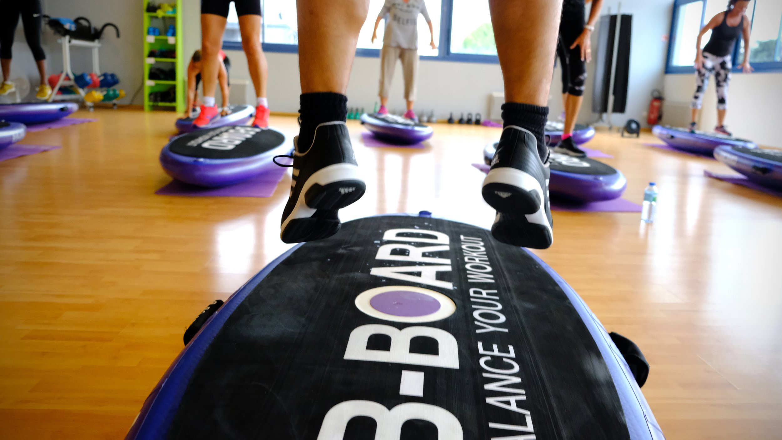 instructor jumping on a balance board B-BOARD® during a fitness class with participants jumping in a fitness room with window in the background