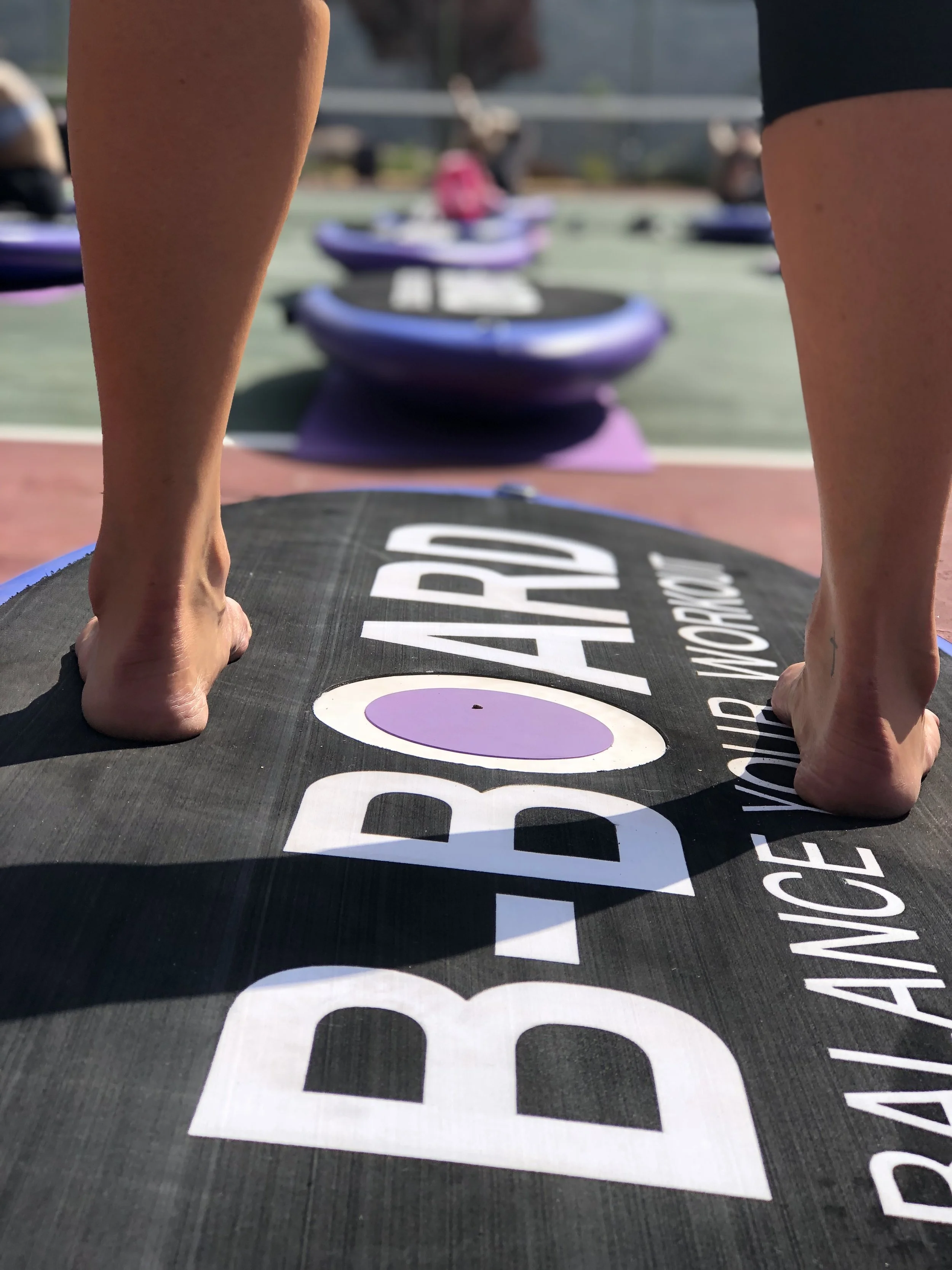 A person standing up on a balance board with the word "B-BOARD" written on it, on an outdoor court with several purple and blue balance trainers in the background.