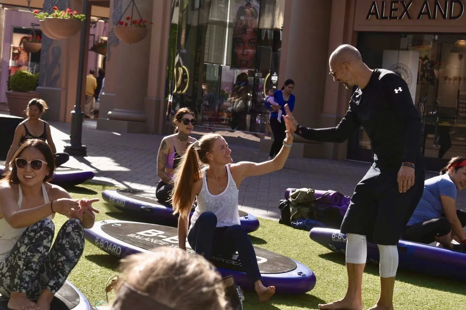 A group of women participating in a balance board class outdoors on artificial grass. An instructor in black sportswear is giving a woman with long red hair a high five, who is sitting on a balance board.