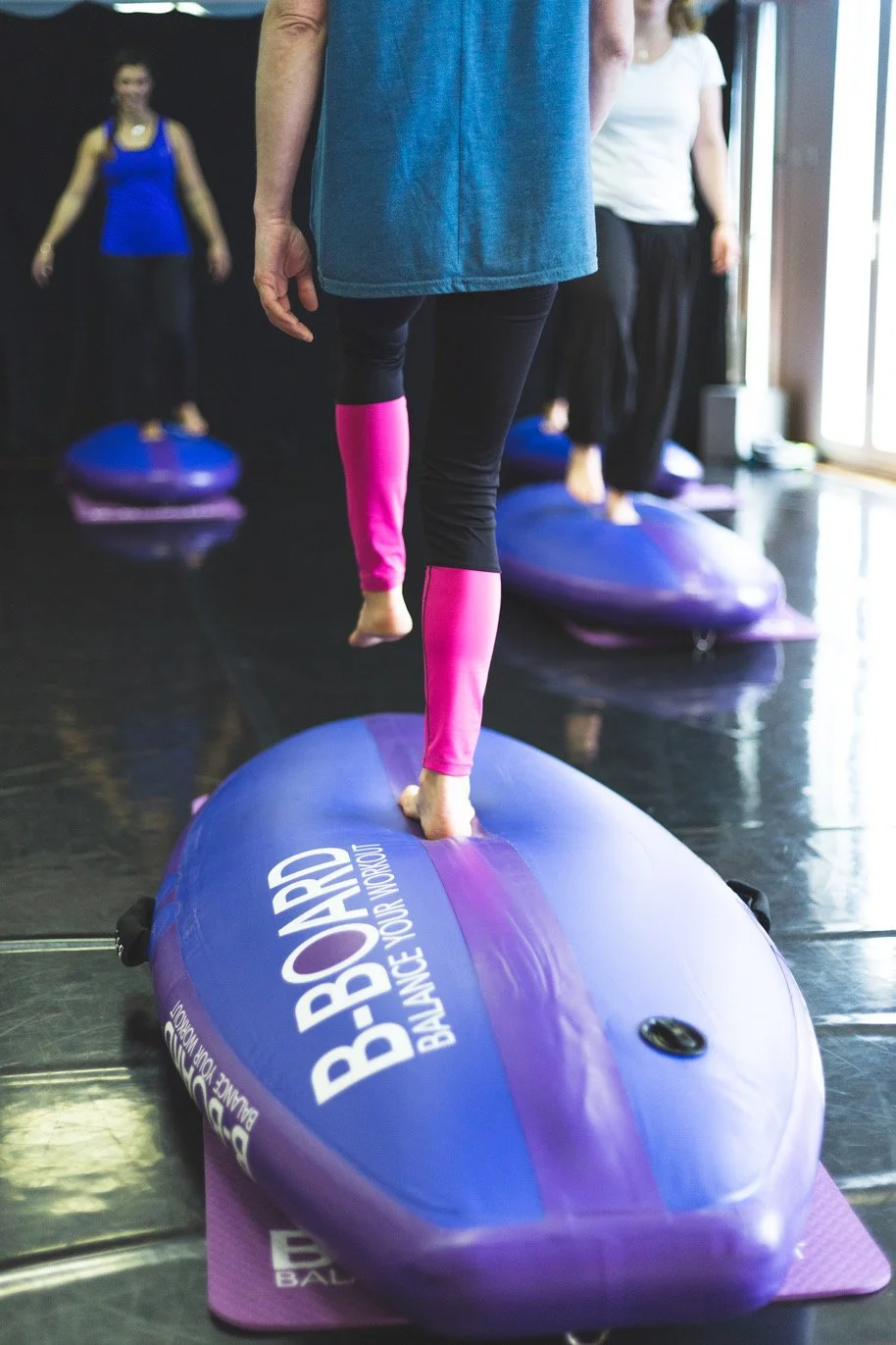 close on an instructor barefoot performing a balance exercise on the round part of a balance board called B-BOARD® with other participants doing the same in the background, in a fitness room