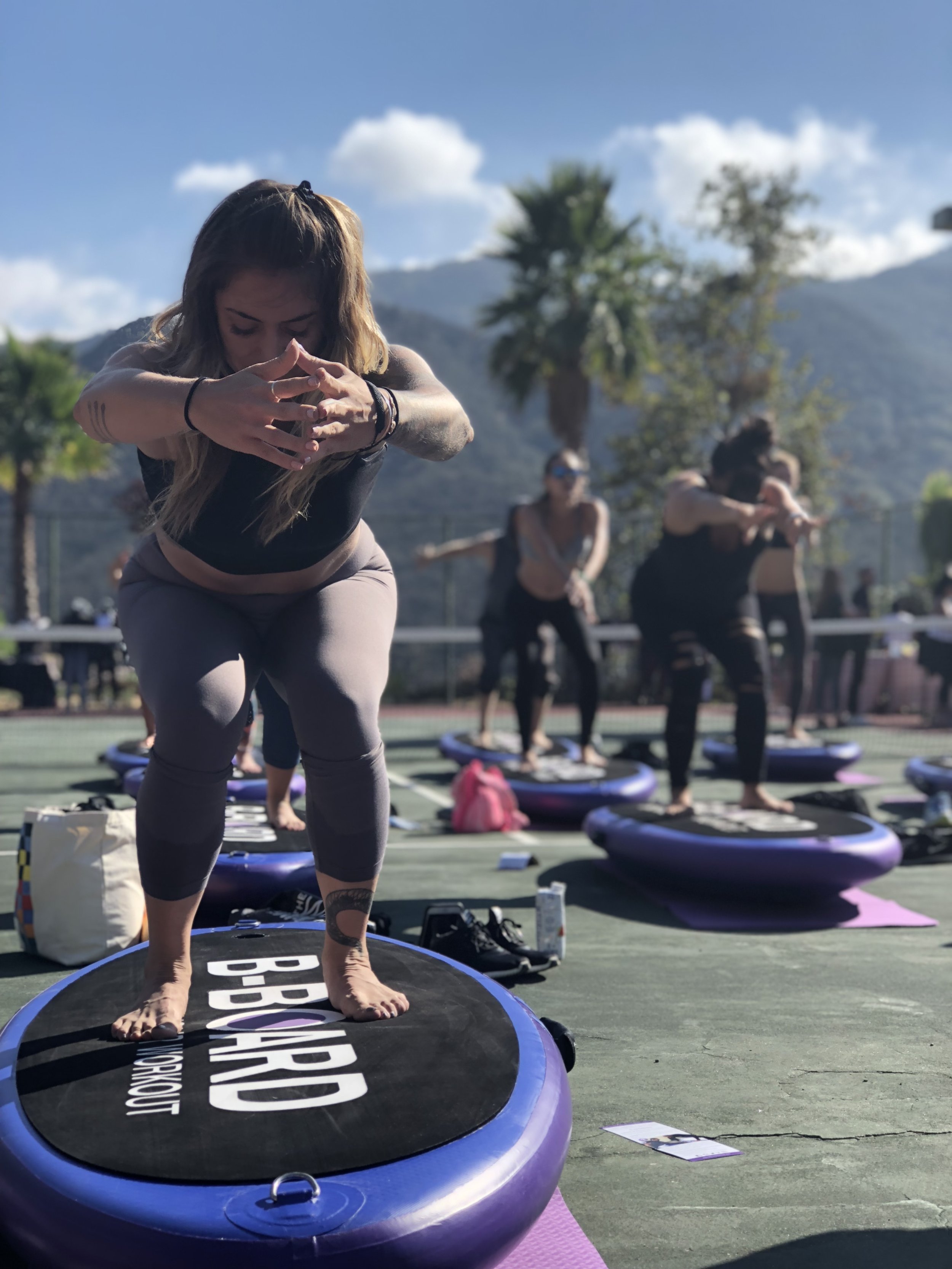Young woman with blue top and black leggins performing a squat on a balance board with B-BOARD on it facing Eric, personal trainer smiling and pointing fingers to encourage his client, in a park setting with green grass and trees in background