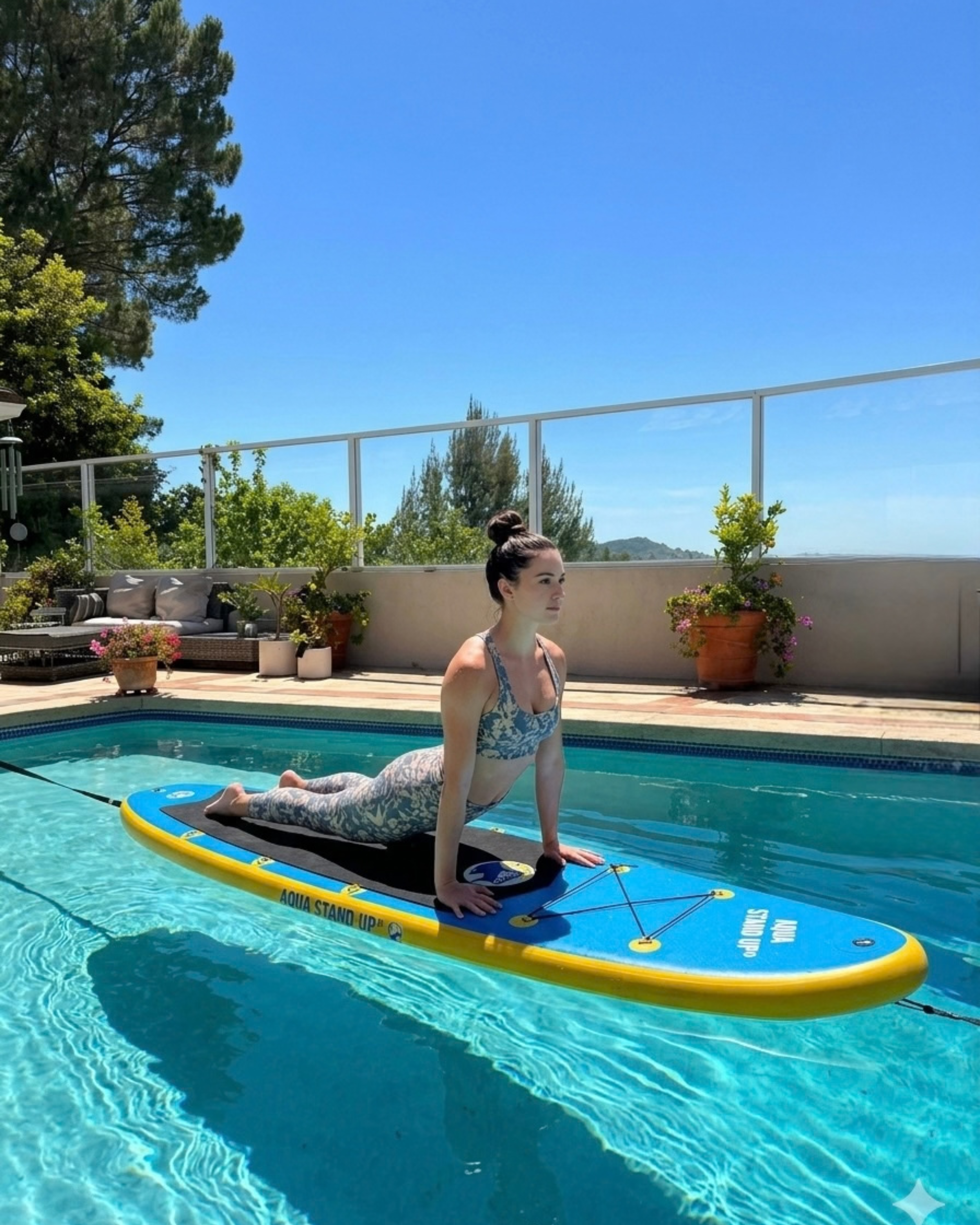 Young woman performing an up dog pose on an AQUA STAND UP® board in her private swimming pool with blue sky and plants in background