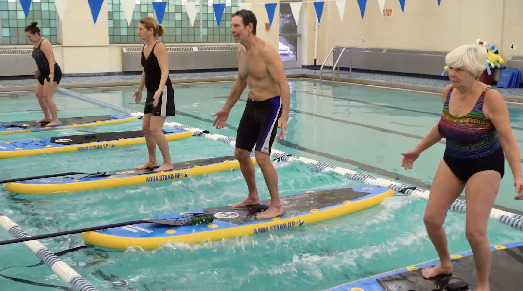 Older adults and young individuals standing on paddleboards on an indoor swimming pool during a class, with an instructor guiding them.