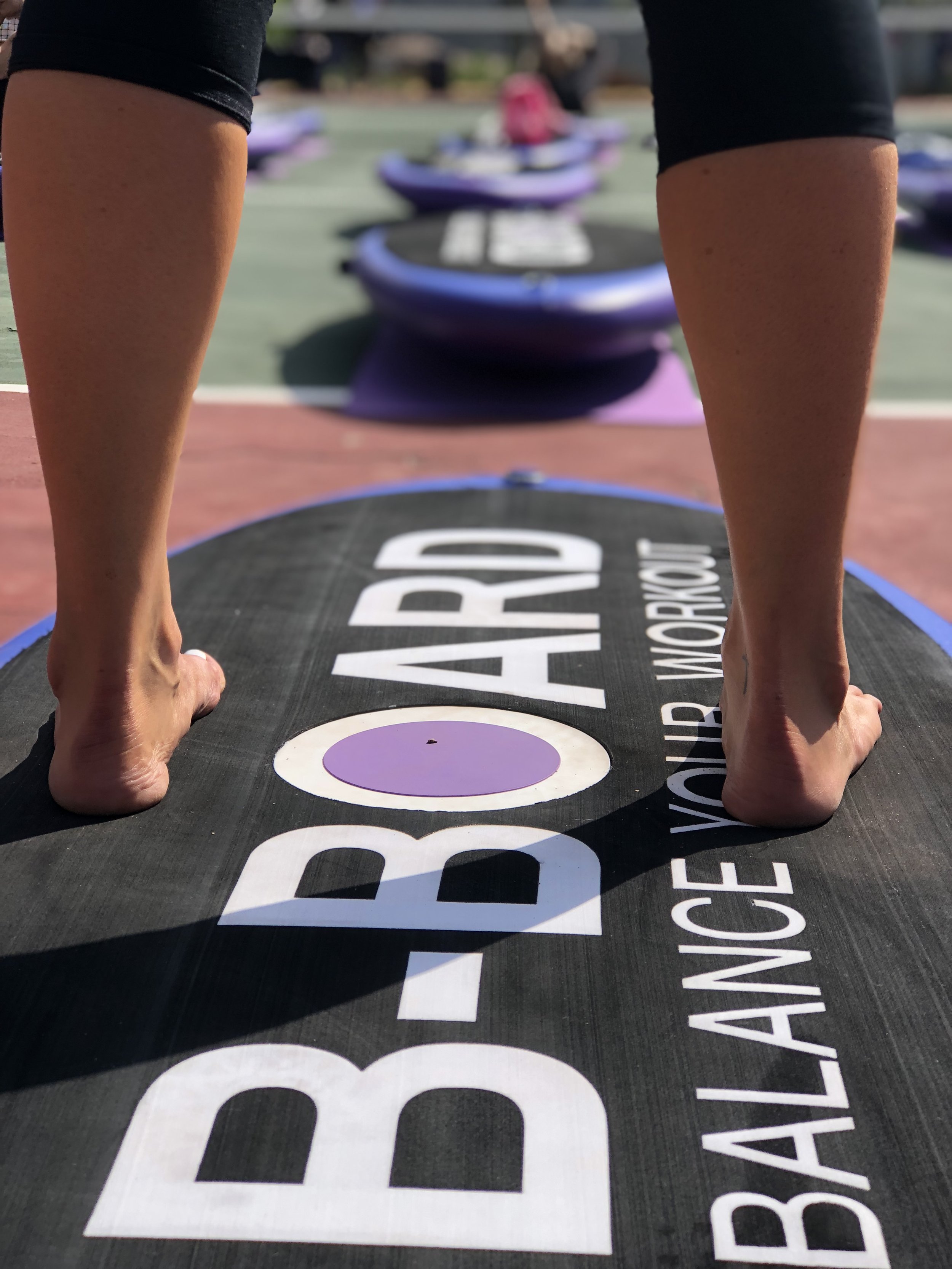 Close-up view of a person standing up on a balance board withh the text b-board balance your workout on the black mat outdoors, with other balance boards and people in the background.