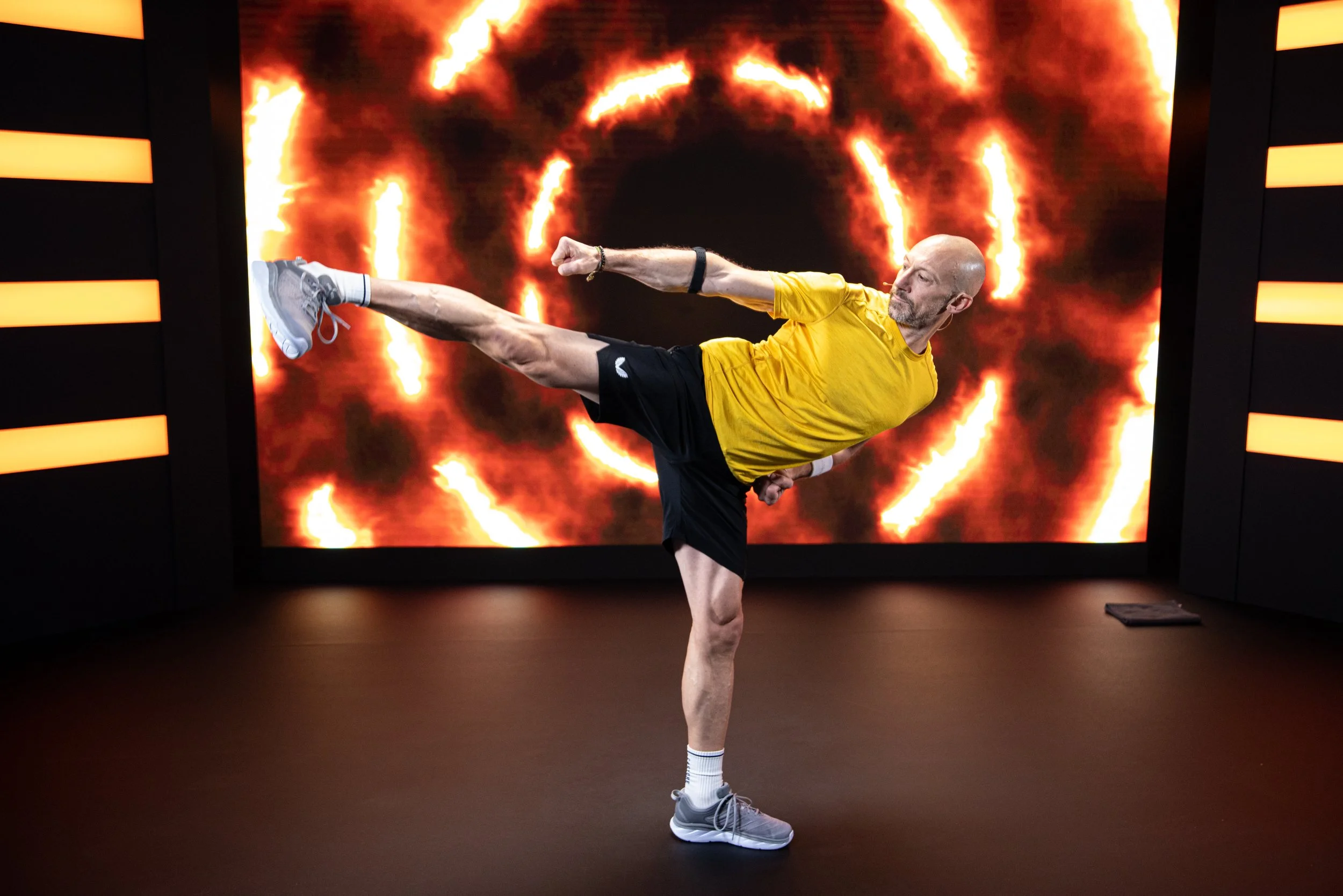 Eric Vandendriessche in a yellow shirt and black shorts performing a high kick at a gym with a fiery digital background.
