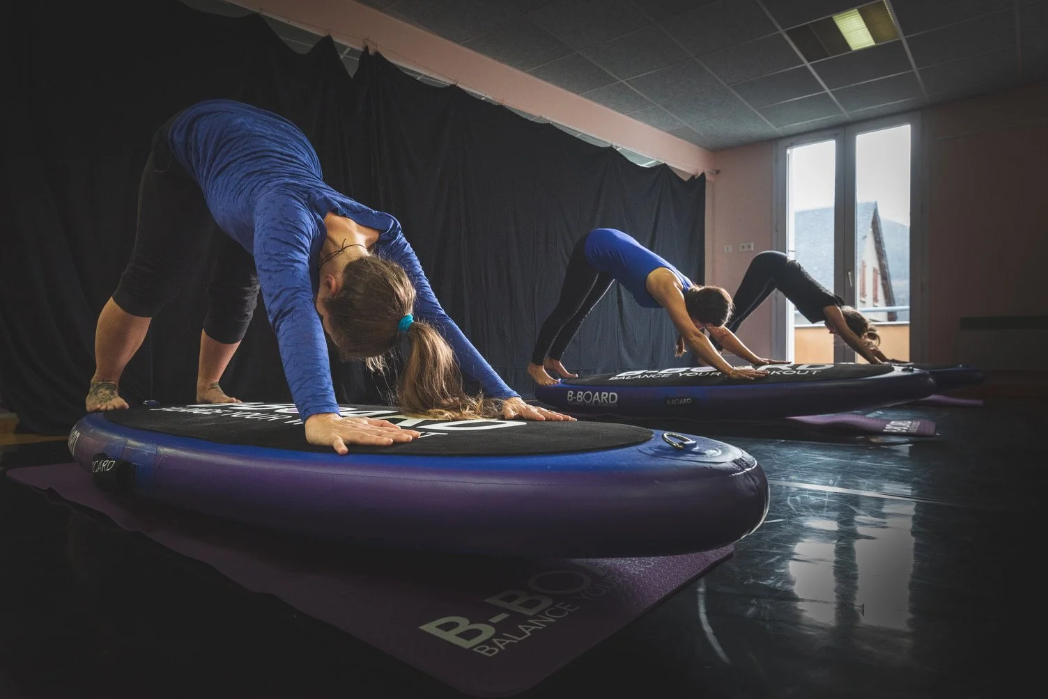 3 young women performing downward Dog, a yoga pose, on a balance board called B-BOARD®, in a fitness room