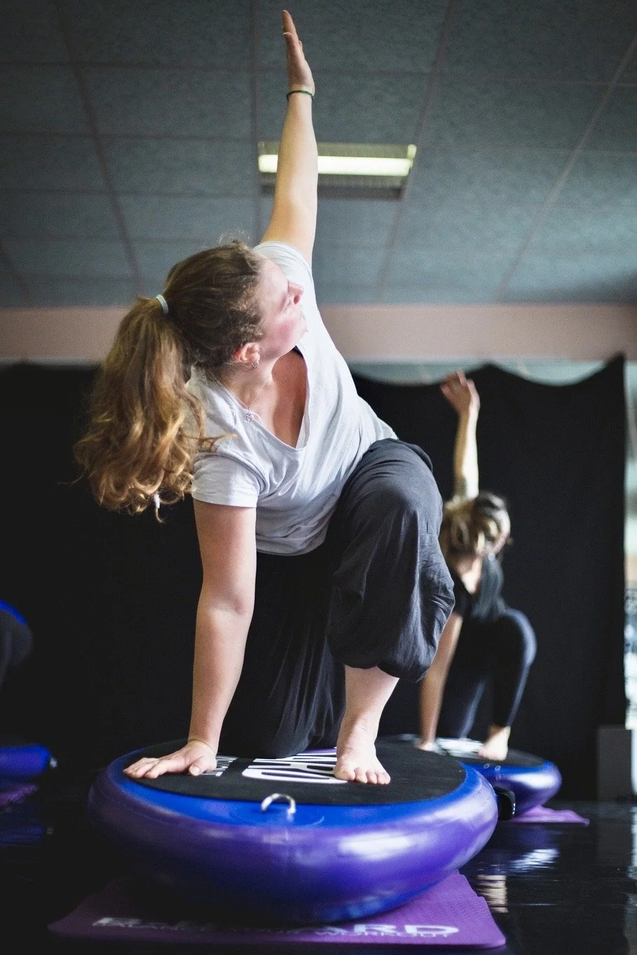 young woman performing a modified side angle pose on a balance board called B-BOARD® with other participants in the background