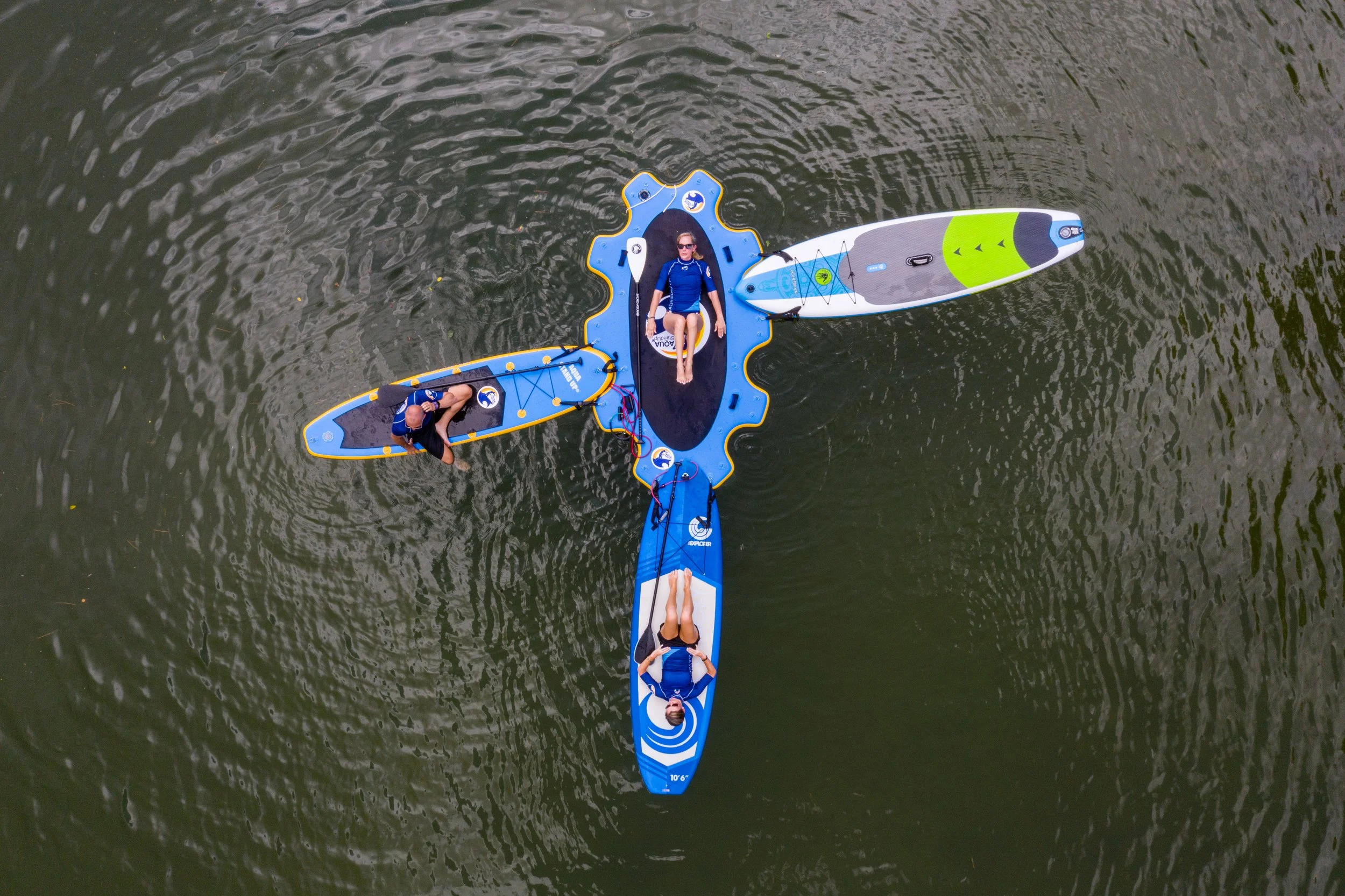 Aerial view of five people on stand-up paddleboards in a body of water, with one person lying on a platform and others sitting or lying on the boards.