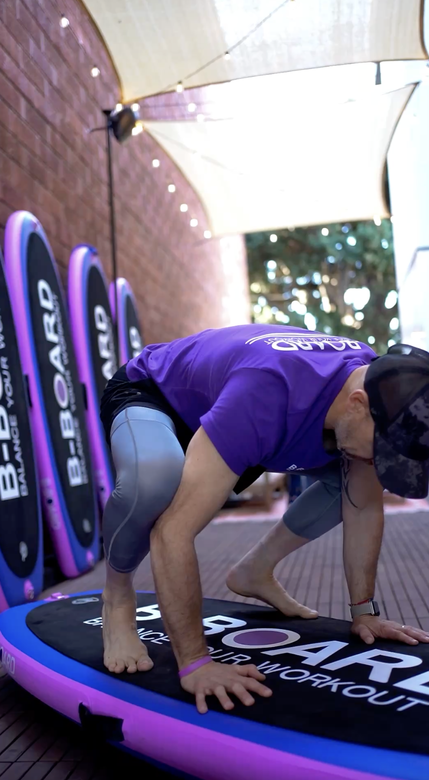 eric vandendriessche, inventor of B-BOARD®, performing a yoga pose called crow pose on a pink, purple and black balance board in a boutique fitness patio with red brick wall, lights and boards against the wall in the background