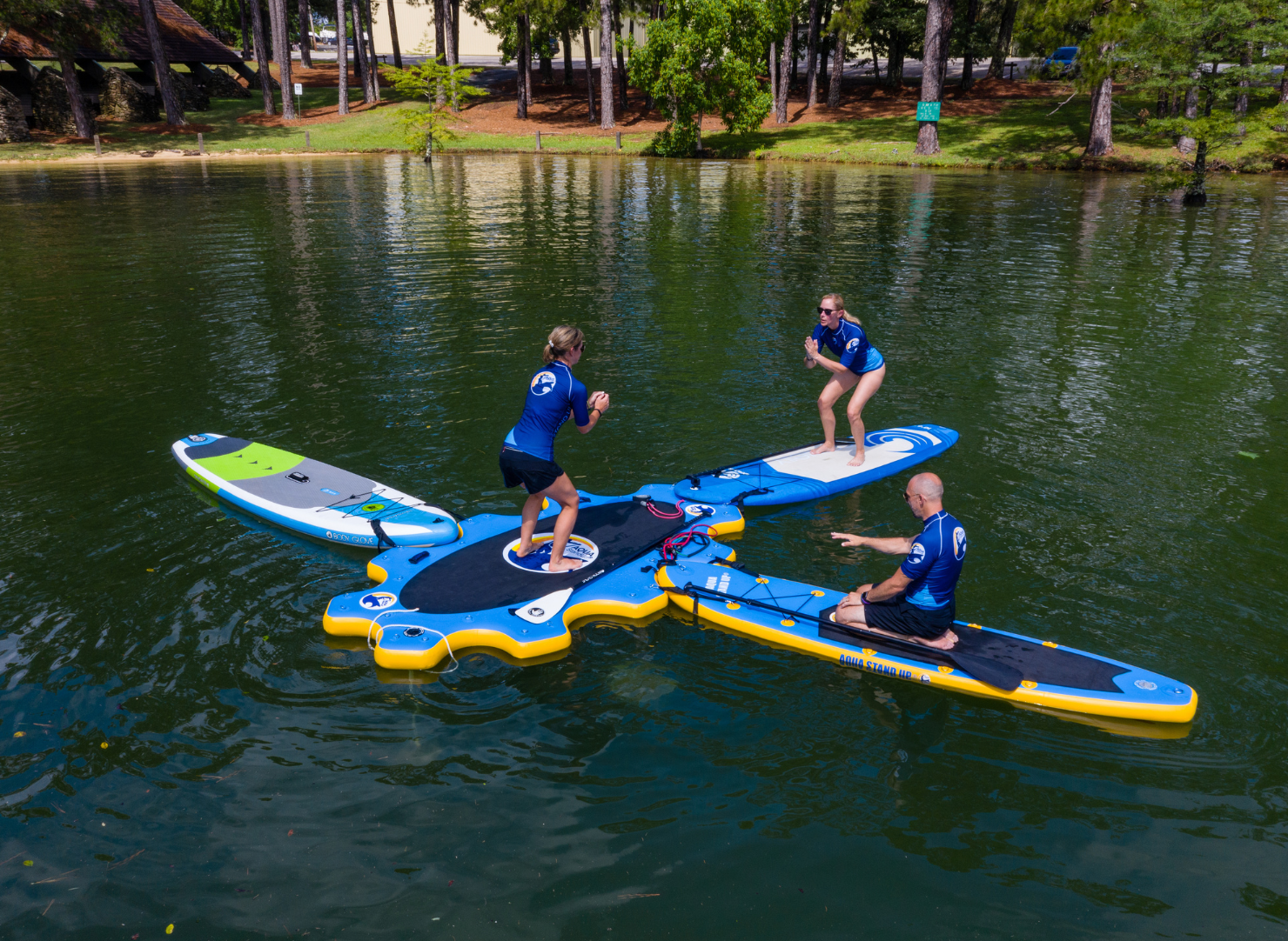 inflatable aquastandup platform set up on a lake with 3 paddle bopards connected to it and people performing squats and a trainer guiding them