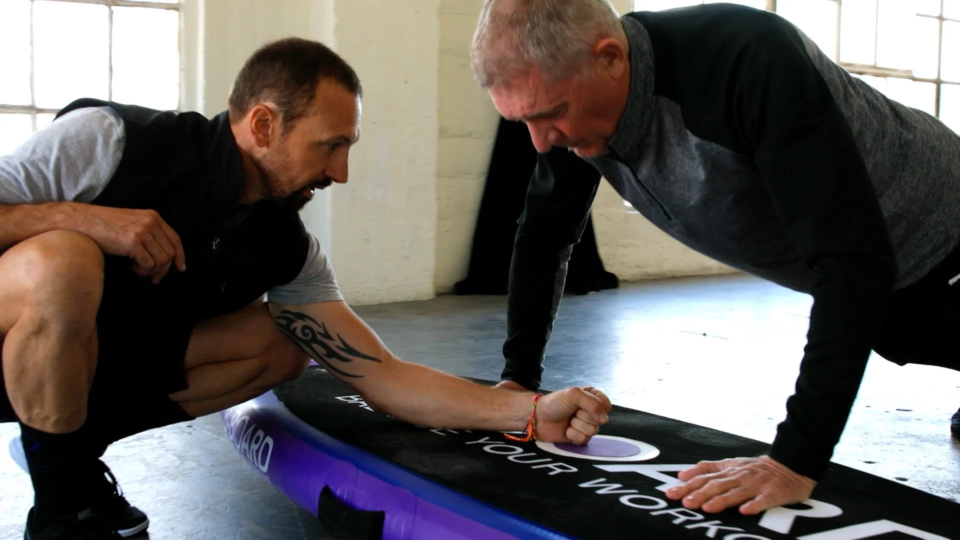 Two men practicing physical exercises on a mat in a bright, industrial-style room with large windows. One man is helping or coaching the other, who is in a plank position.