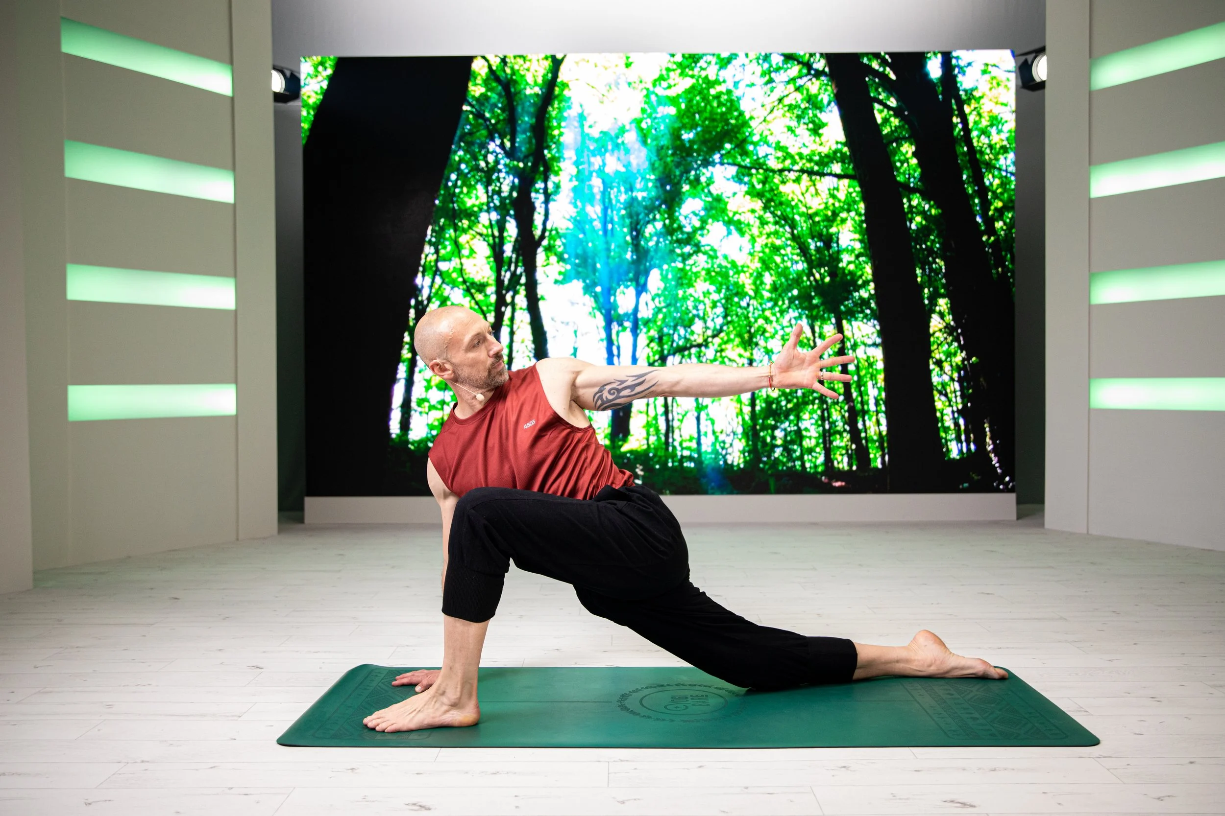 Eric Vandendriessche practicing yoga on a green mat, performing a low lunge pose with one arm extended forward and the other resting on his knee, in a room with a large digital display of a green forest background.