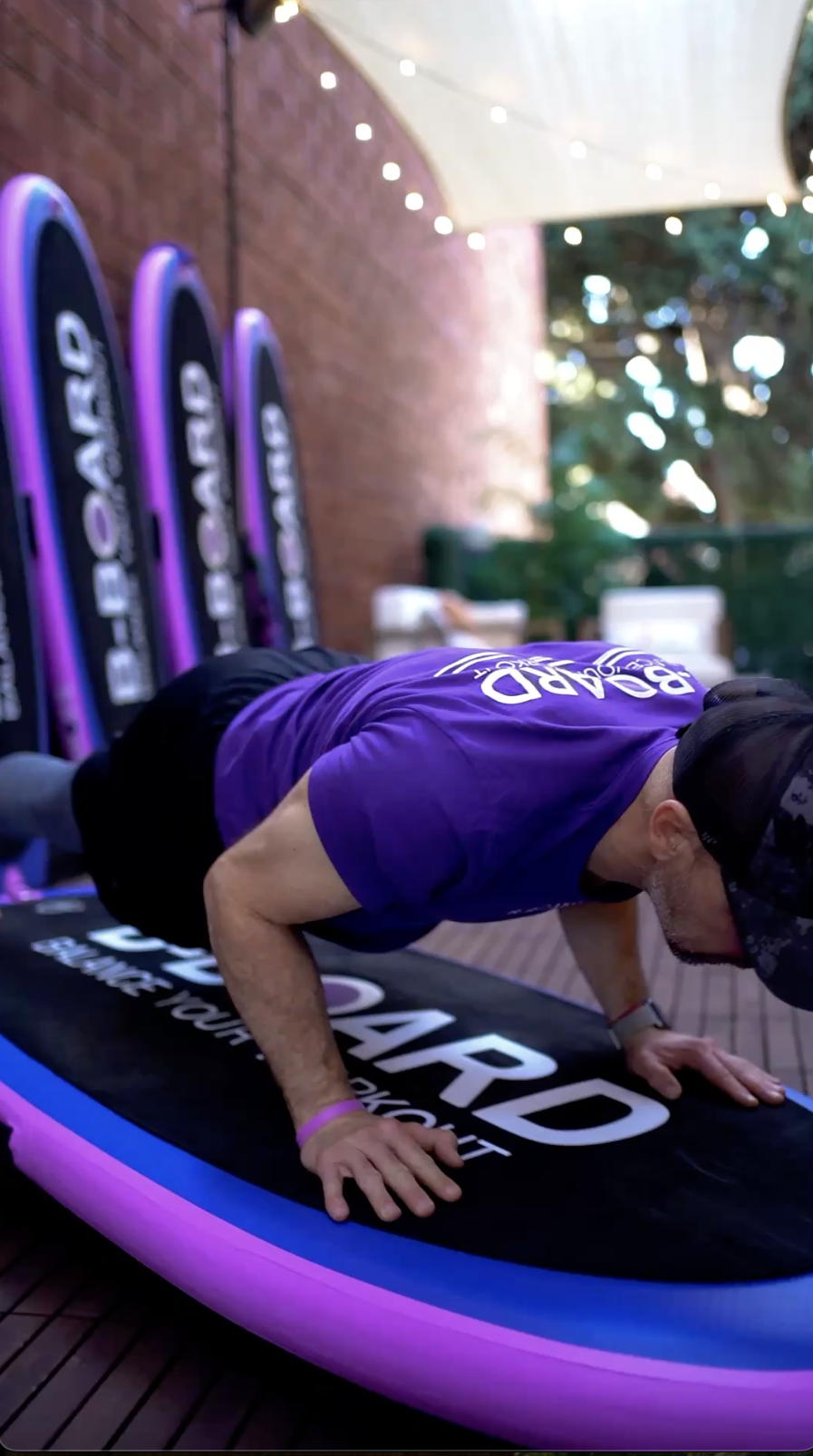 eric vandendriessche, inventor of B-BOARD®, performing a yoga combination on a pink, purple and black balance board in a boutique fitness patio with red brick wall, lights and boards against the wall in the background