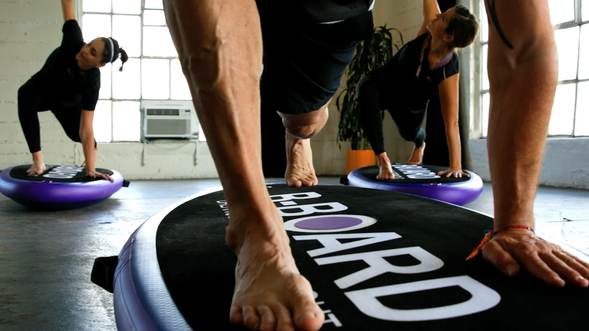 People practicing yoga on balance board B-BOARD® in a studio with large windows.