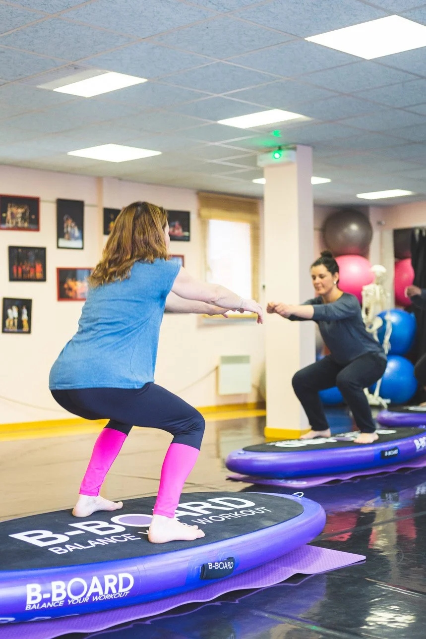female instructor performing a squat on a balance board called B-BOARD® facing female student performing a squat on her board, in a fitness room with window and swiss balls in the background