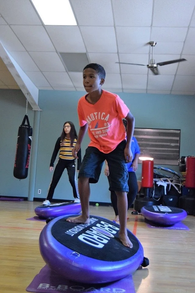 A young boy is balancing on a purple balance board called B-BOARD® in a fitness studio, with other children practicing exercises in the background during a B-BOARD® Kids class .