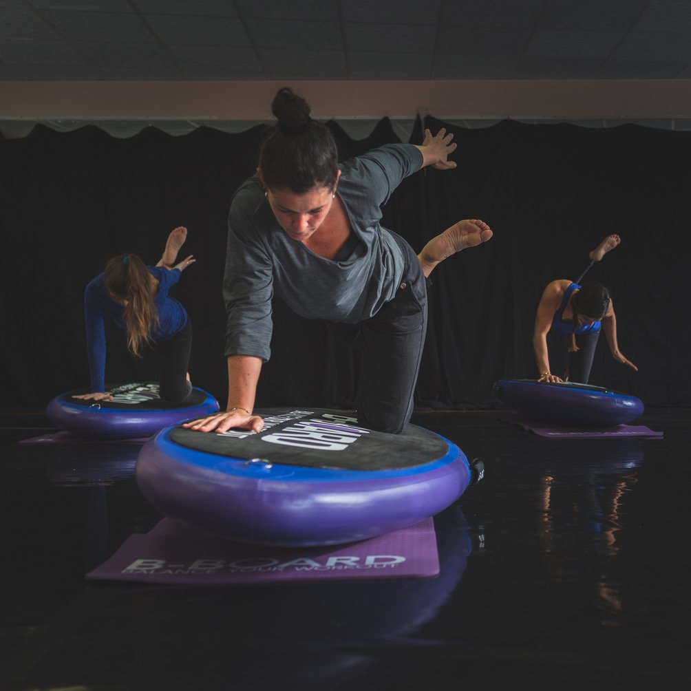 young woman on a balance board performing a half bow pose, a yoga pose with participants in the background