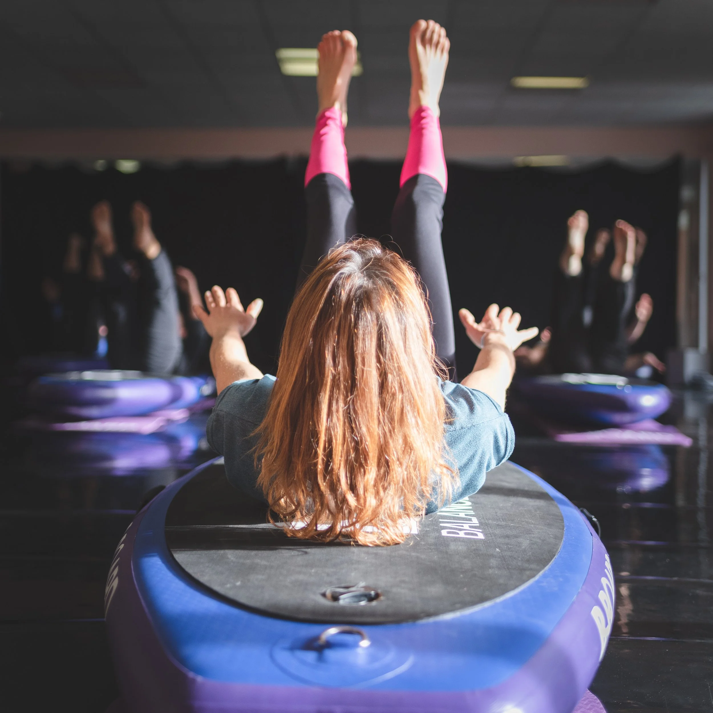 female instructor in front of participants during a B-BOARD® yoga class, performing a 100 Pilates on a balance board, in a fitness room