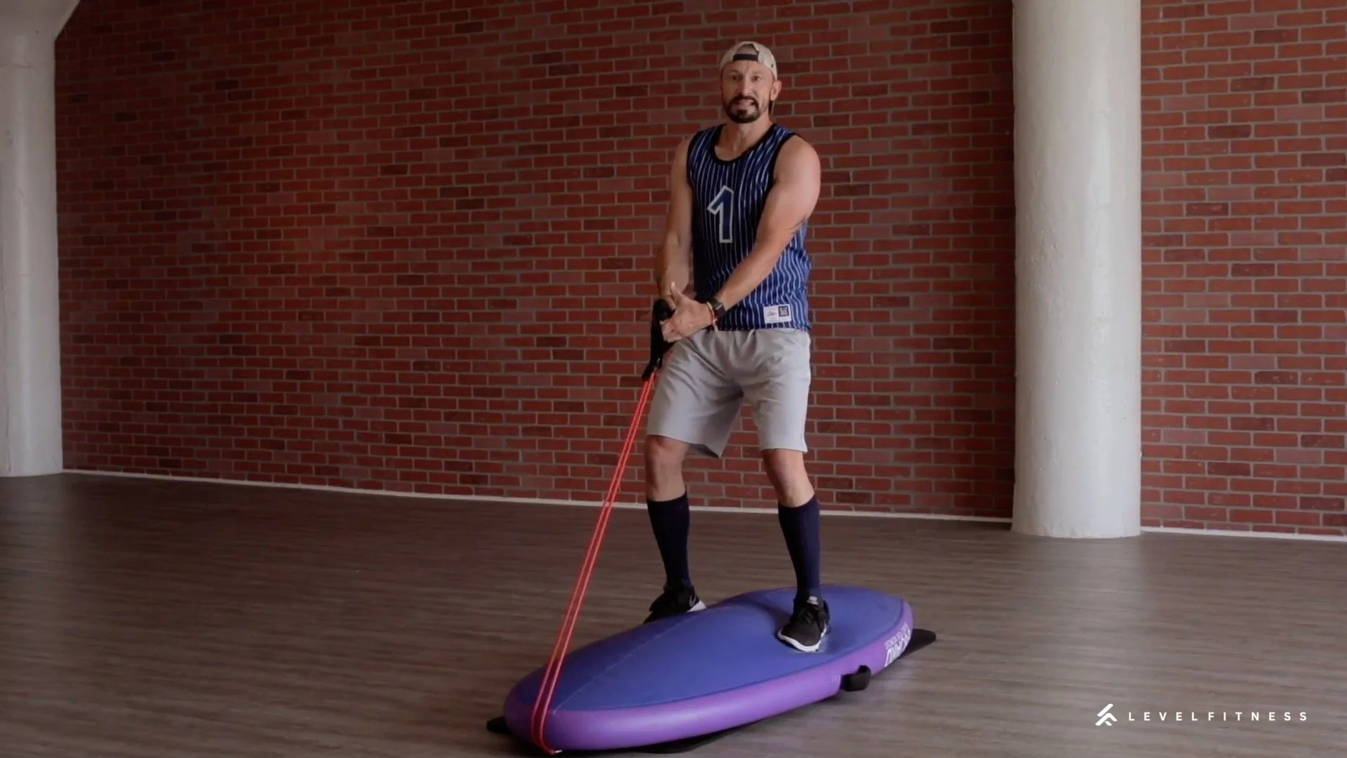 A man balancing on a purple balance board flipped over while holding a resistance band in a gym setting with a brick wall background.