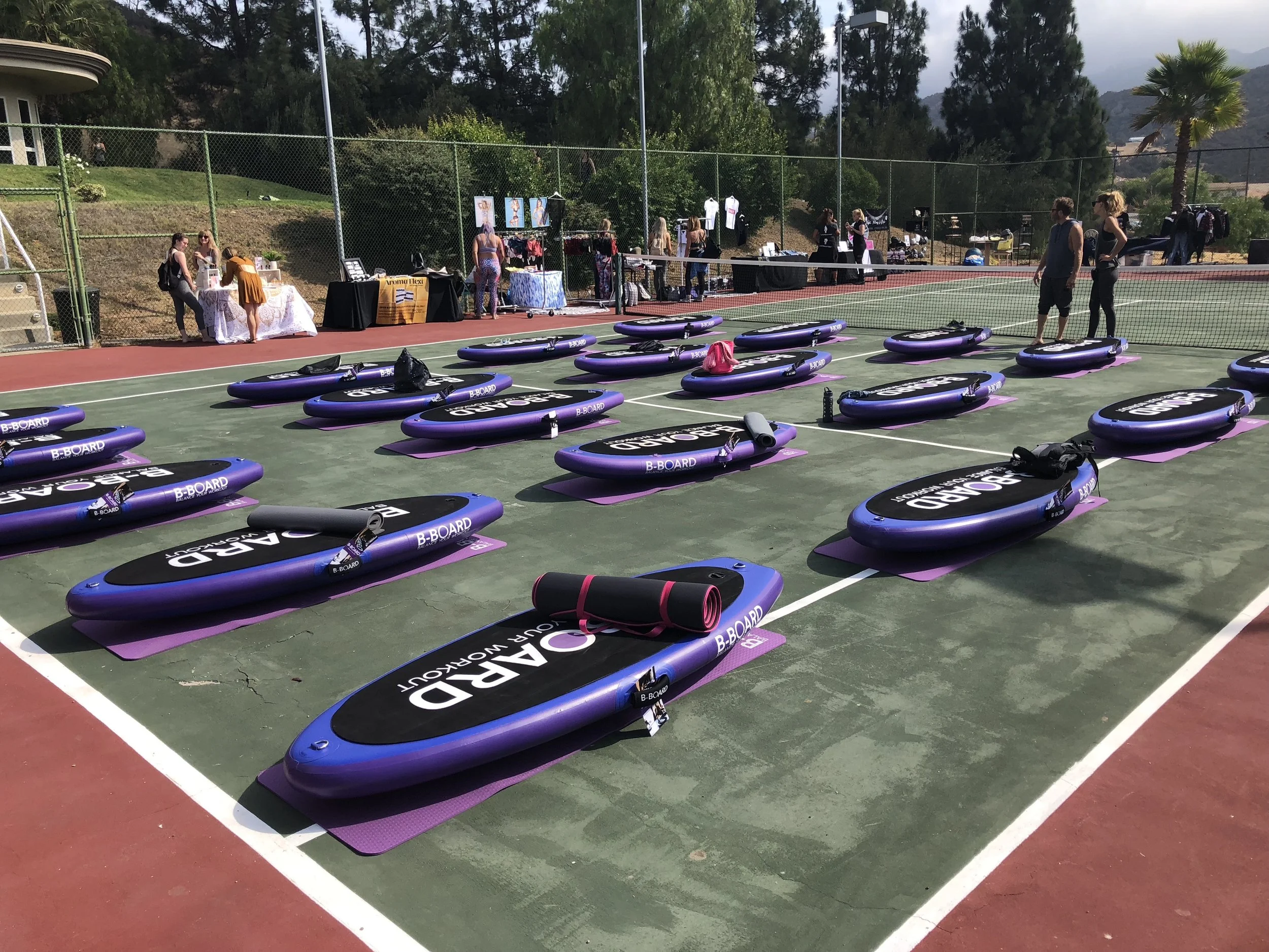 A tennis court repurposed for a yoga class, with multiple purple and black inflatable balance boards arranged on the court. Several people are setting up various booths along the fence in the background, with trees and mountains visible behind them.
