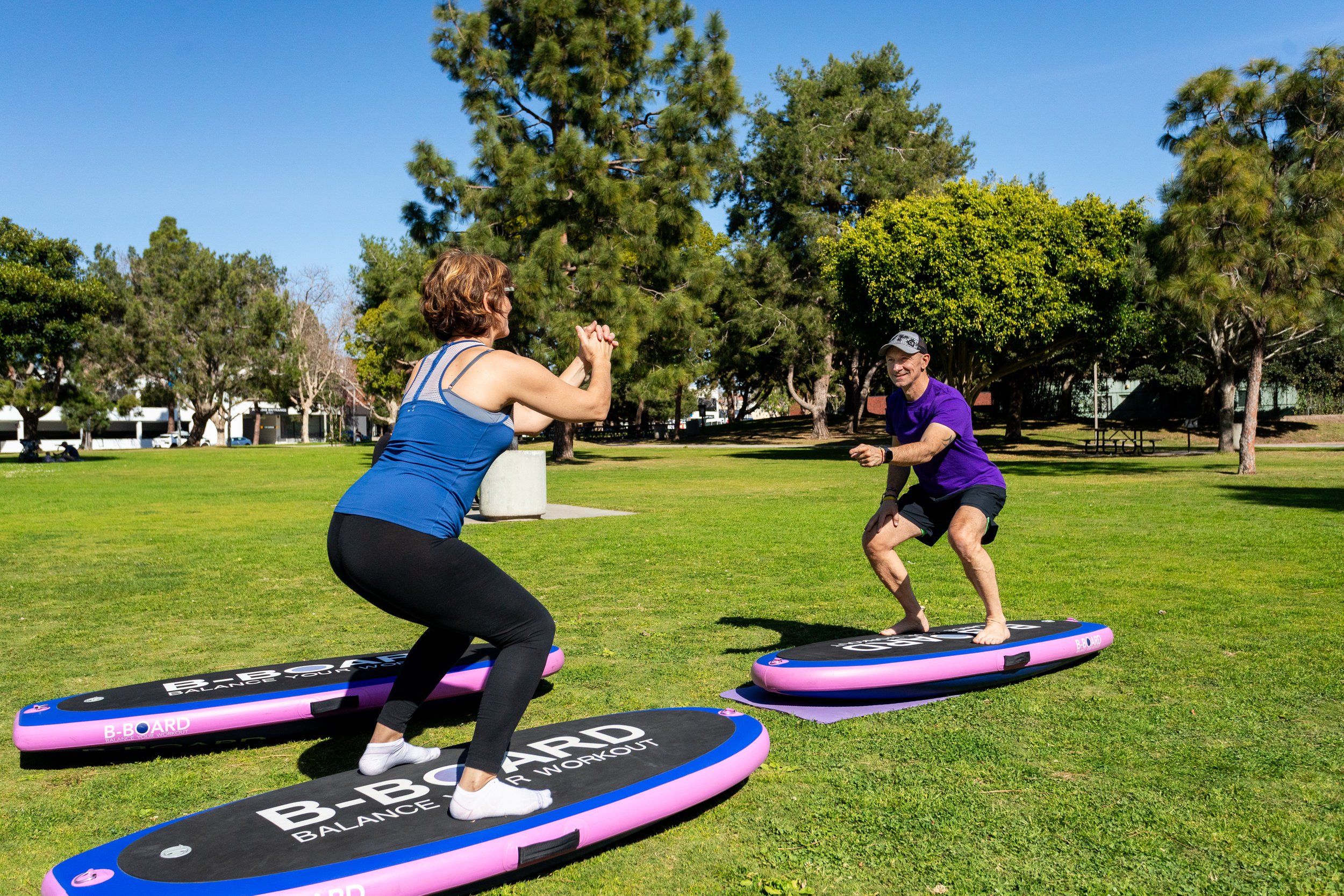 Young woman with blue top and black leggins performing a squat on a balance board with B-BOARD on it facing Eric, personal trainer smiling and pointing fingers to encourage his client, in a park setting with green grass and trees in background