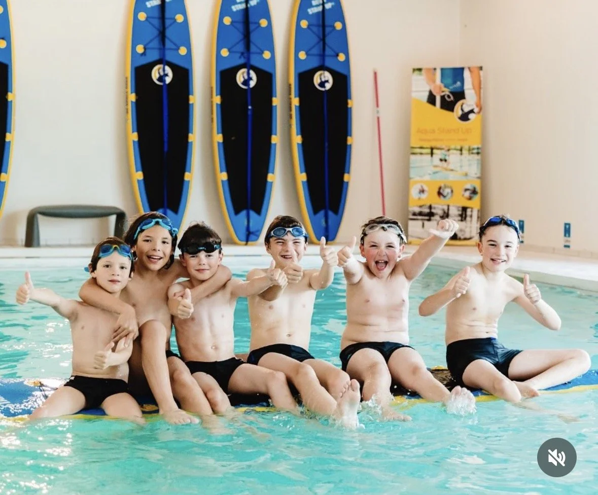Group of smiling children sitting on a yellow aqua stand up board in a swimming pool, wearing swim goggles and black swim trunks, giving thumbs up.