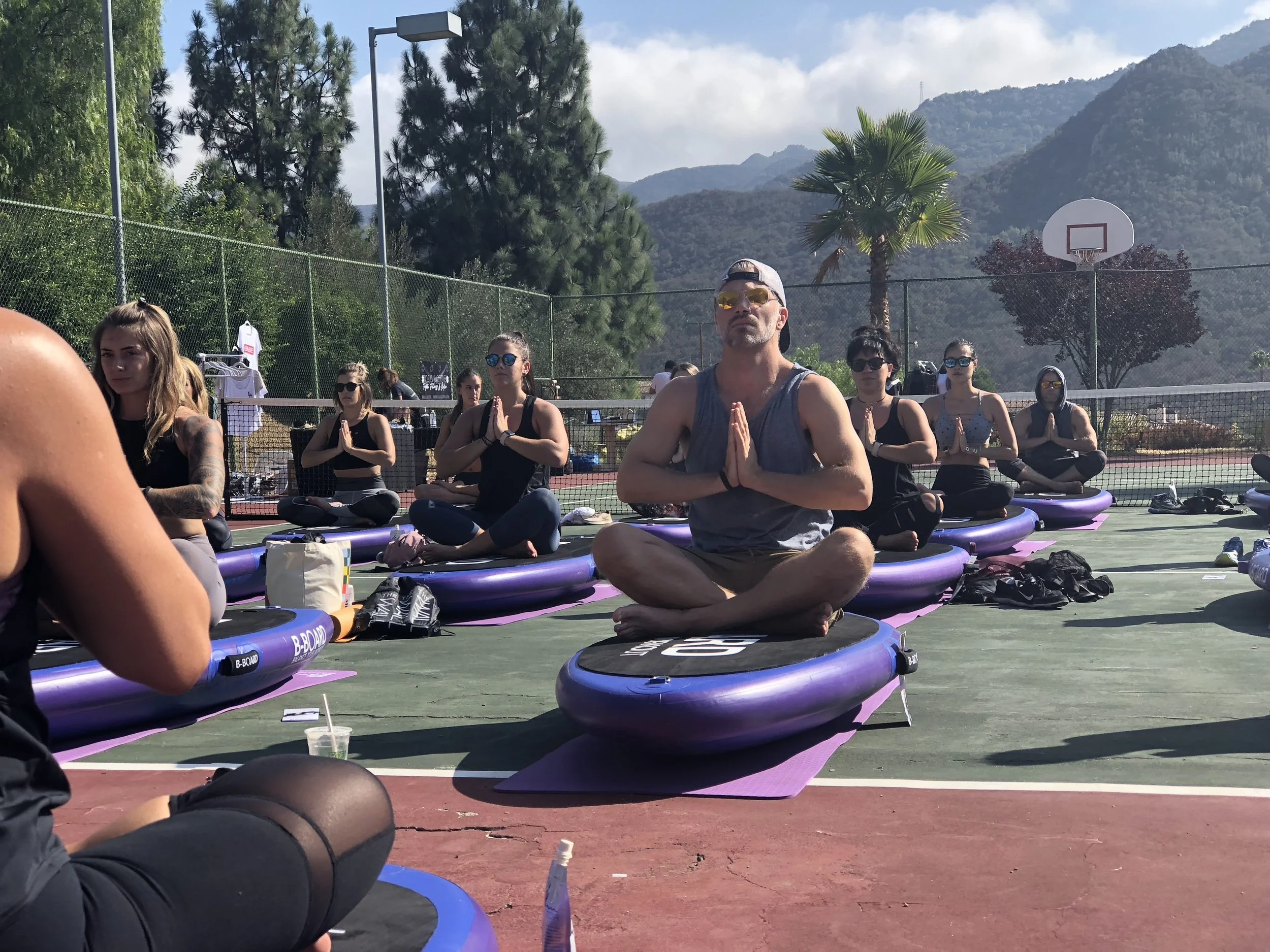 People participating in an outdoor bboard yoga session on a tennis court, seated on purple balance boards with hands in a prayer position, surrounded by trees and mountains in the background.
