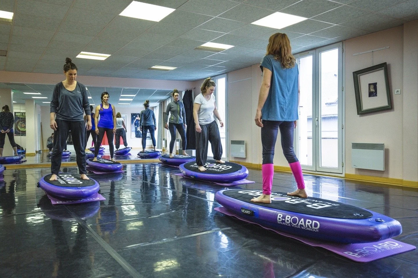 group of young women facing a female instructor during a B-BOARD® yoga class on balance board in a fitness room with windows