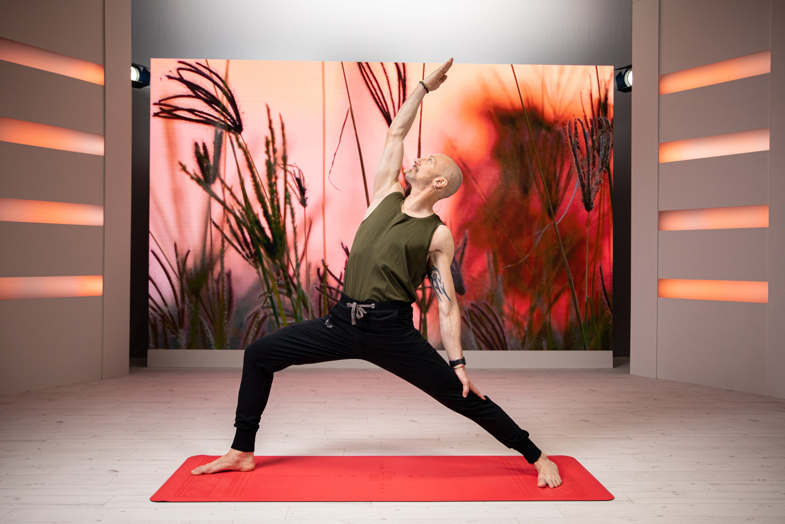Eric Vandendriessche practicing yoga in a studio, performing a reverse warrior pose on a red yoga mat, with a backdrop of tall grasses and a pink-orange sunset