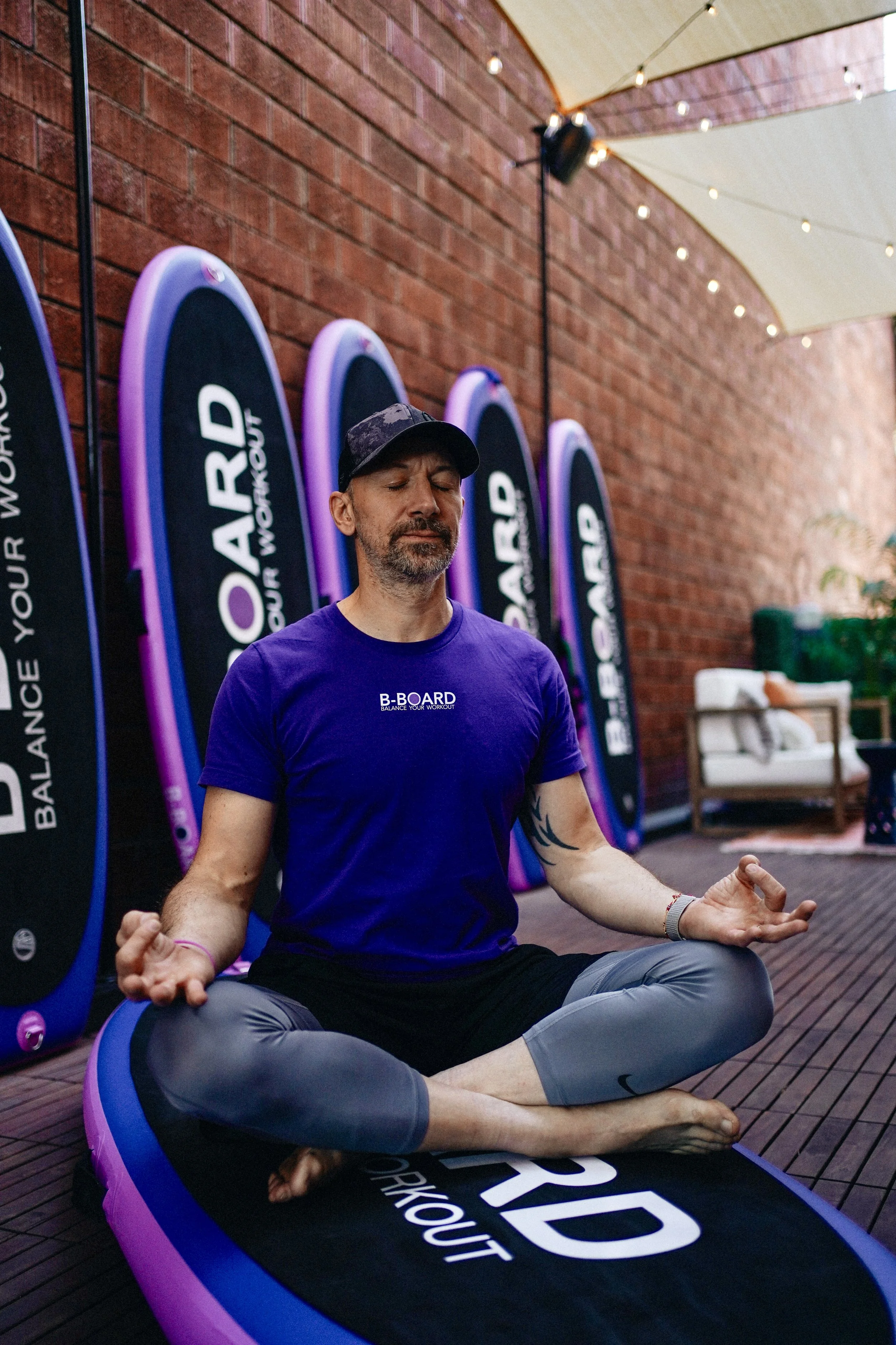 eric vandenriessche, inventor of B-BOARD®, meditating on a pink, purple and black balance board, in a patio with red brick wall, string lights and furniture in the background