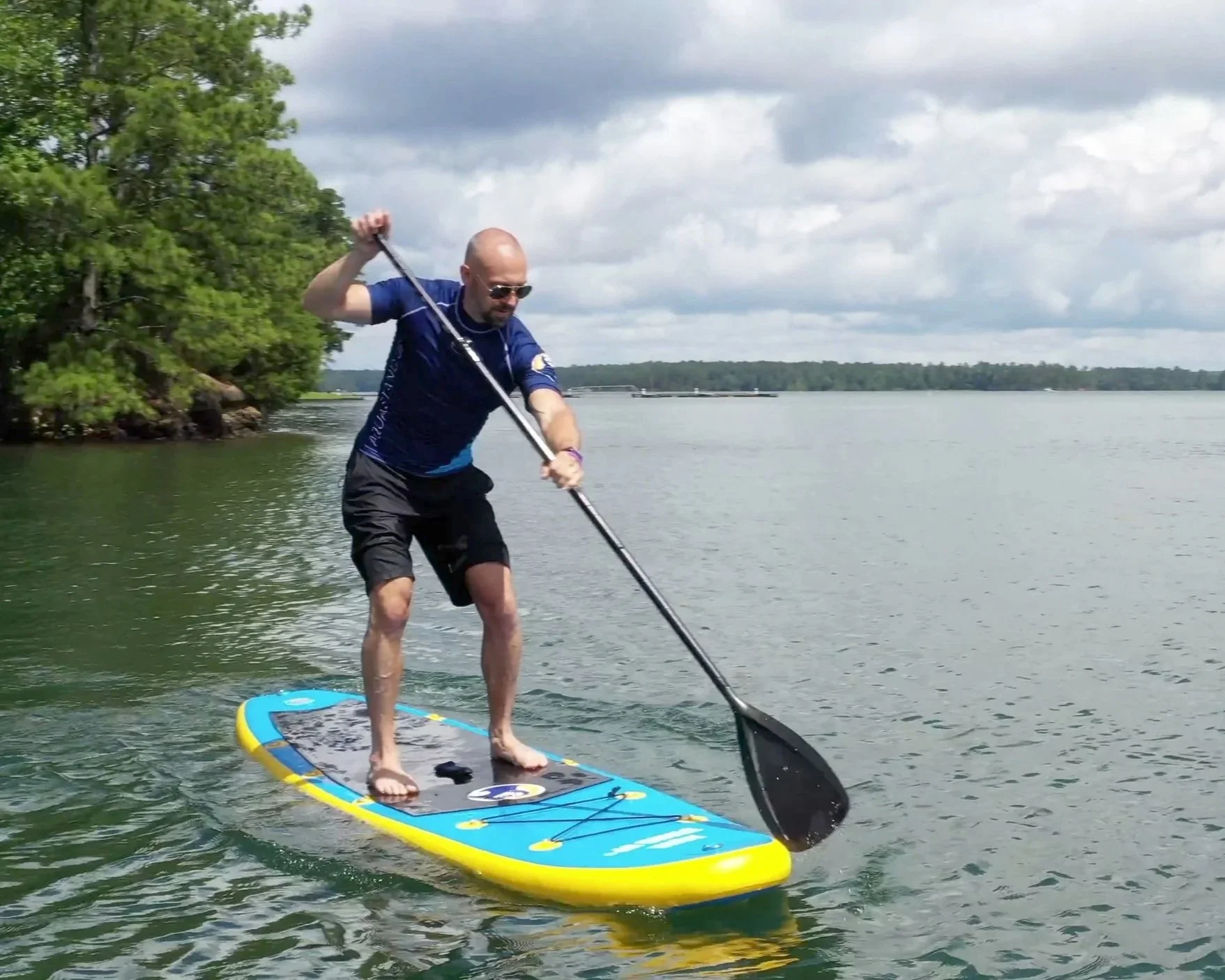 A man paddleboarding on a body of water under a cloudy sky, wearing sunglasses, a blue shirt, and black shorts.