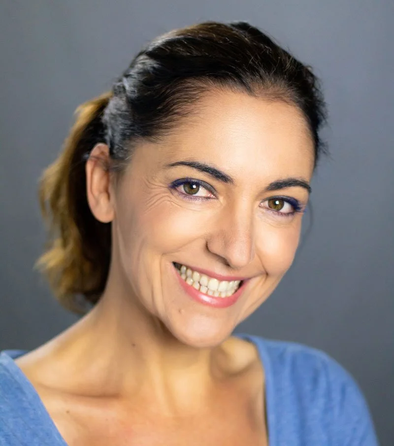 Smiling woman with brown hair wearing a blue top against a gray background.