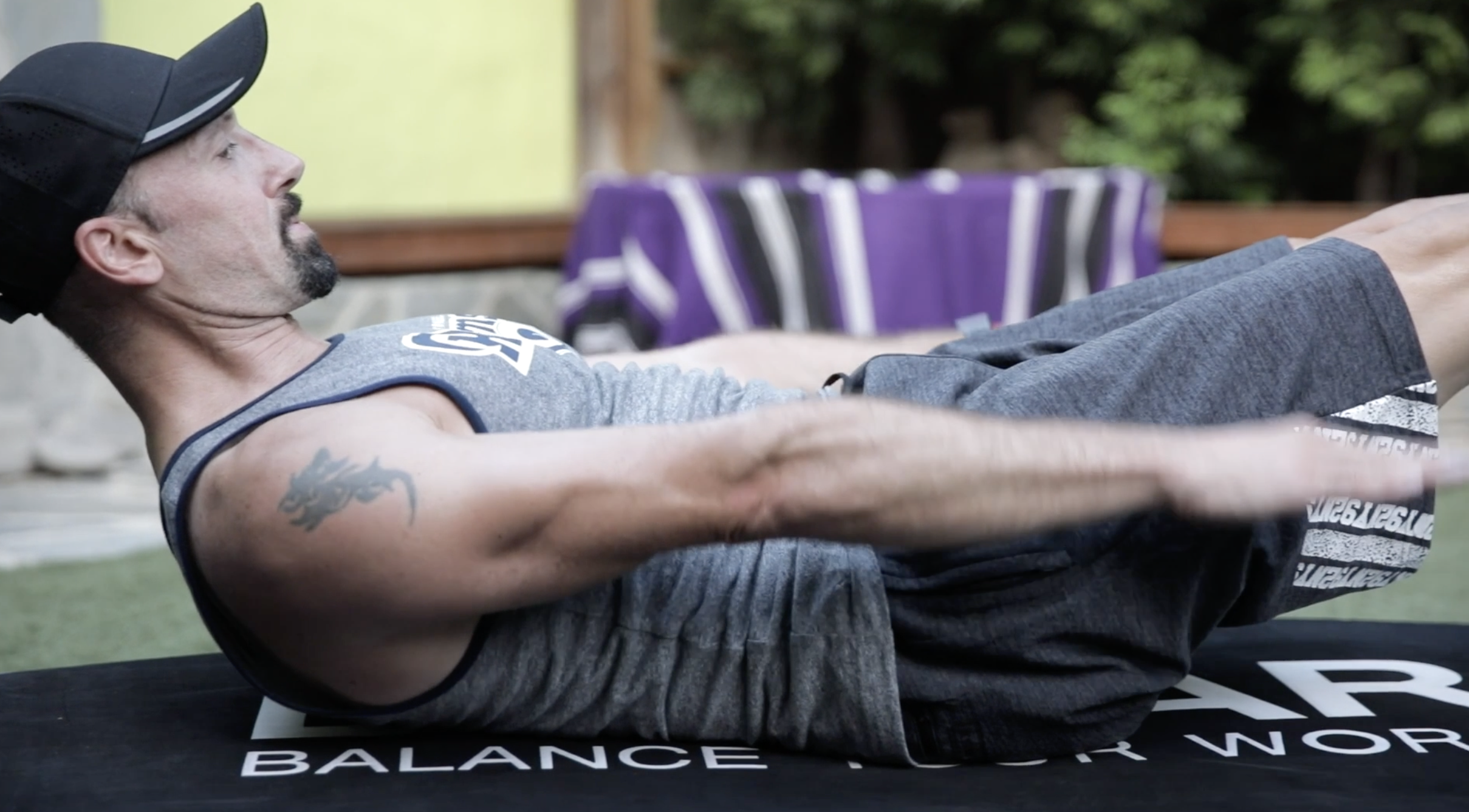 Man lying on a balance board performing a 100 Pilates exercise outdoors, wearing a gray tank top, gray shorts, a black cap, and sporting a tattoo on his left arm.