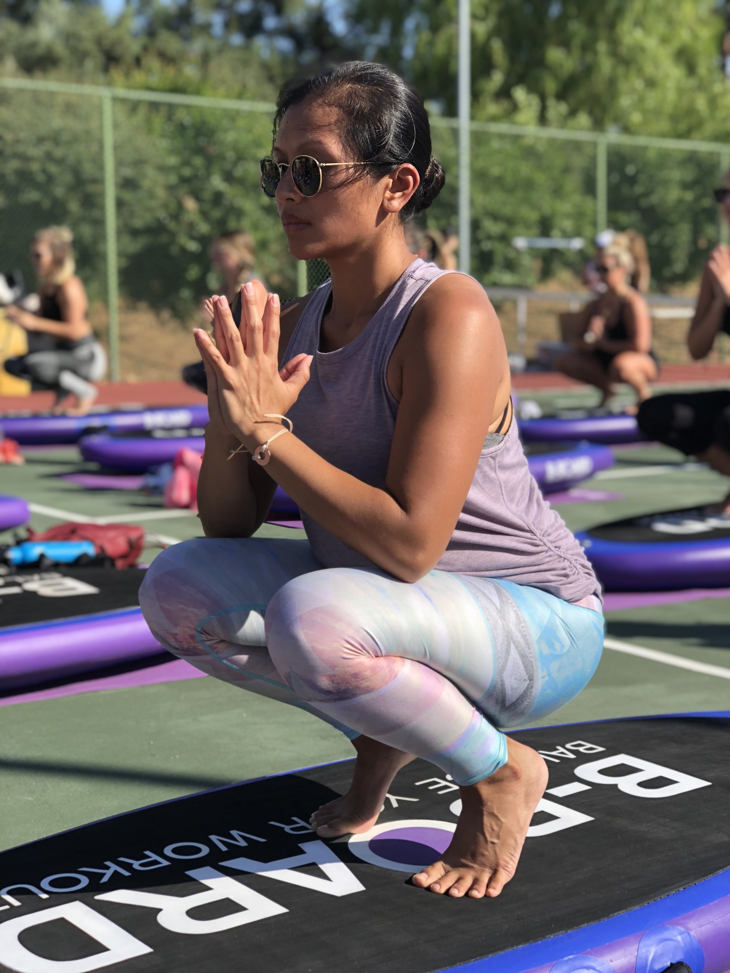 Woman practicing in an outdoor yoga on b-boards, balancing in a squat with hands pressed together in prayer position, wearing sunglasses and colorful leggings.