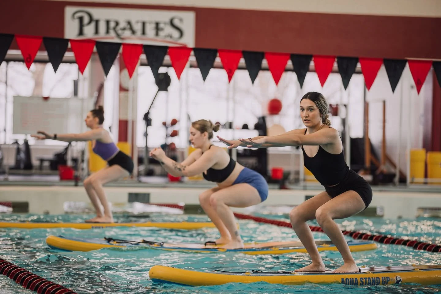 young women in swimsuit performing squats on aqua stand up paddle boards in a swimming pool with PIRATES written on a banner