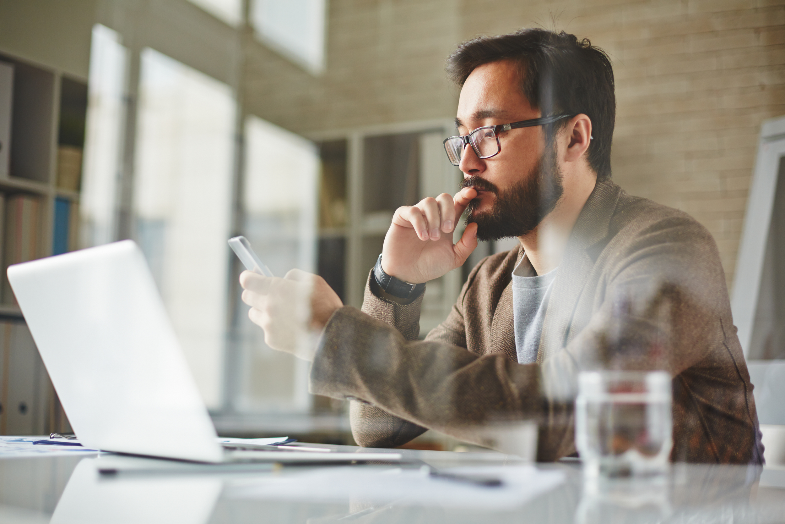 A busy professional holding a smartphone in front of his computer in his office