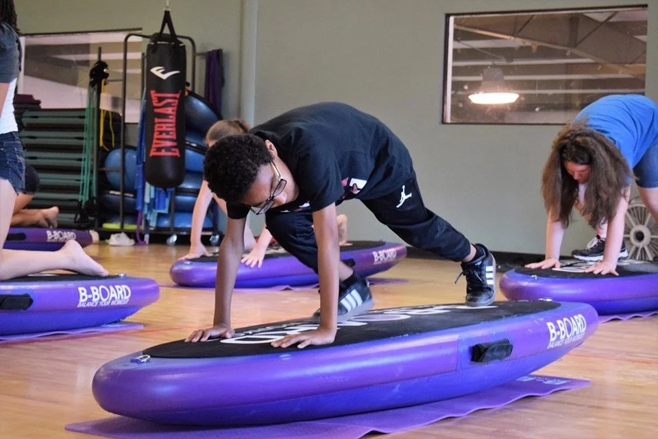 Children participating in a B-BOARD® fitness class, doing plank exercises on purple B-Board balance boards in a gym.