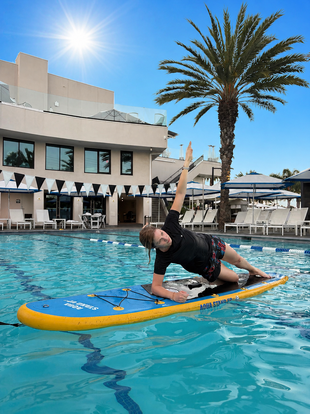 Woman performing a lateral plank on an aqua stand up paddle board in the swimming pool at the griffin club los angeles with sunny blue sky and palm tree