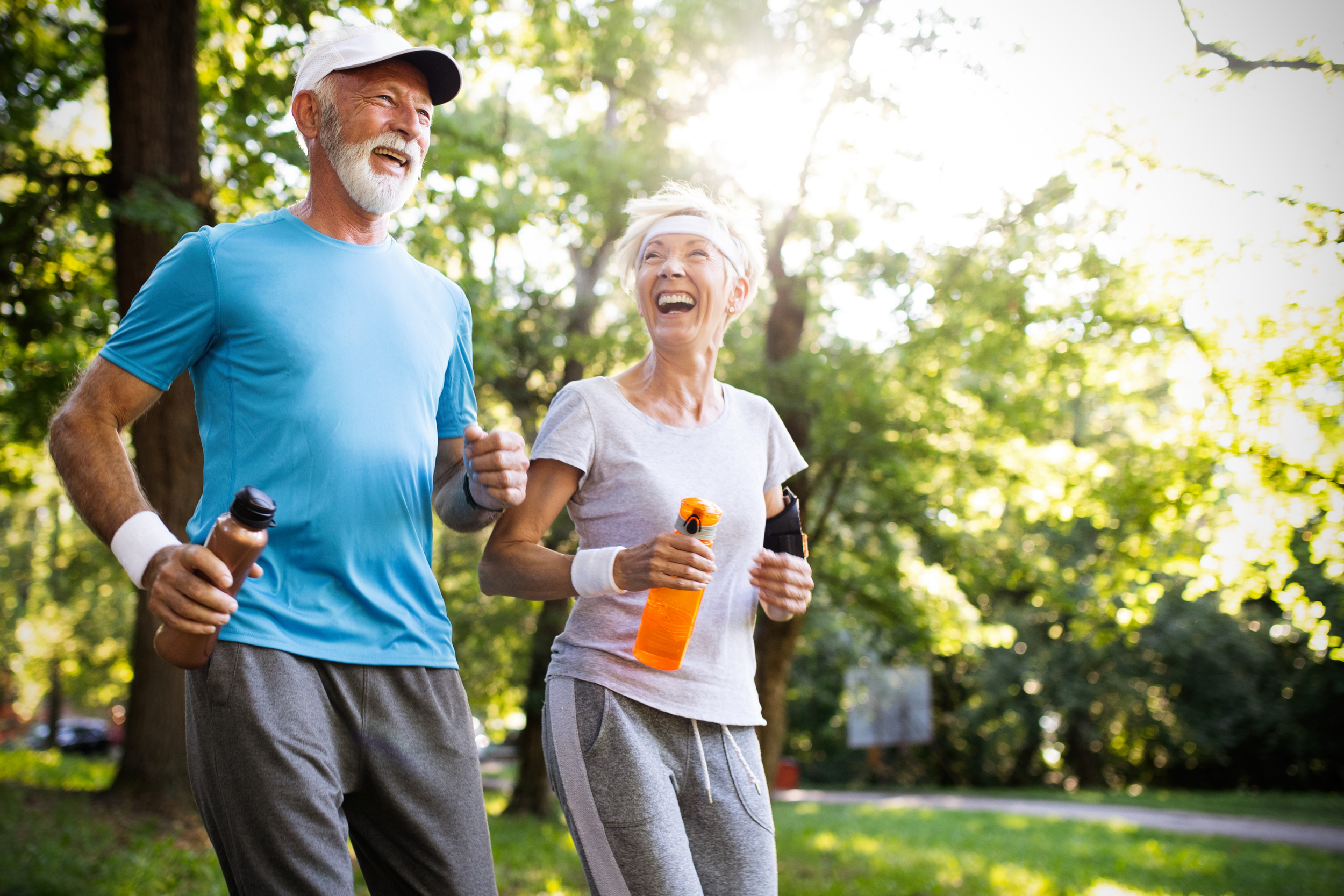 a couple of active agers smiling and jogging surrounded by trees and grass on a sunny day