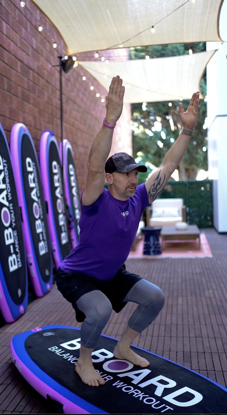 eric vandendriessche, inventor of B-BOARD®, performing a yoga pose called chair pose with arms extended on a pink, purple and black balance board in a boutique fitness patio with red brick wall, lights and boards against the wall in the background