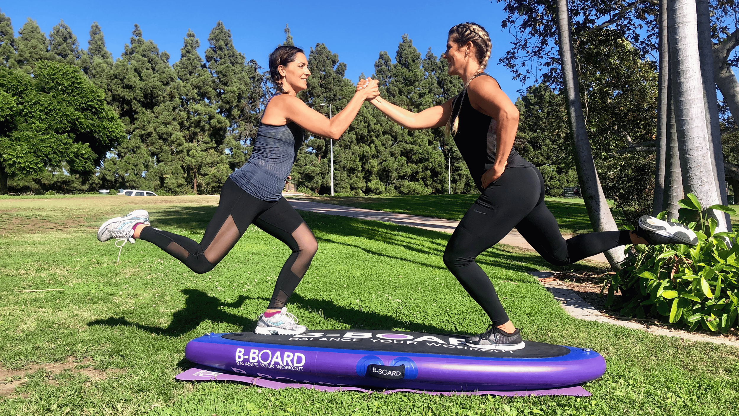 2 young women holding their hands on a balance board called B-BOARD® in a park with green grass and trees, and performing a squat on 1 leg