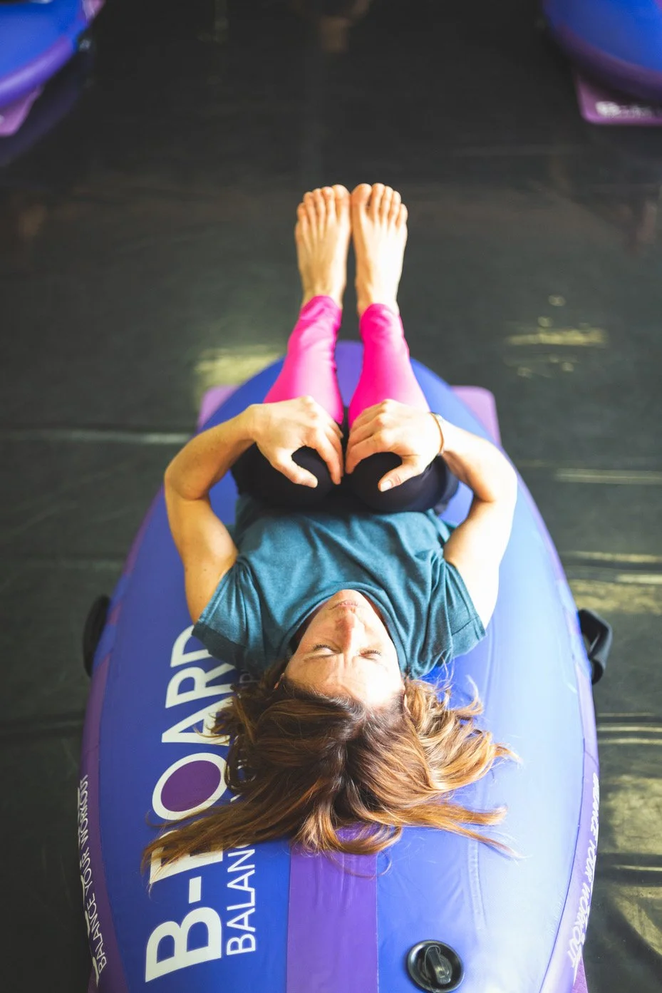 female instructor holding her knees to her chest during a yoga class on balance board called B-BOARD®, with her board flipped over