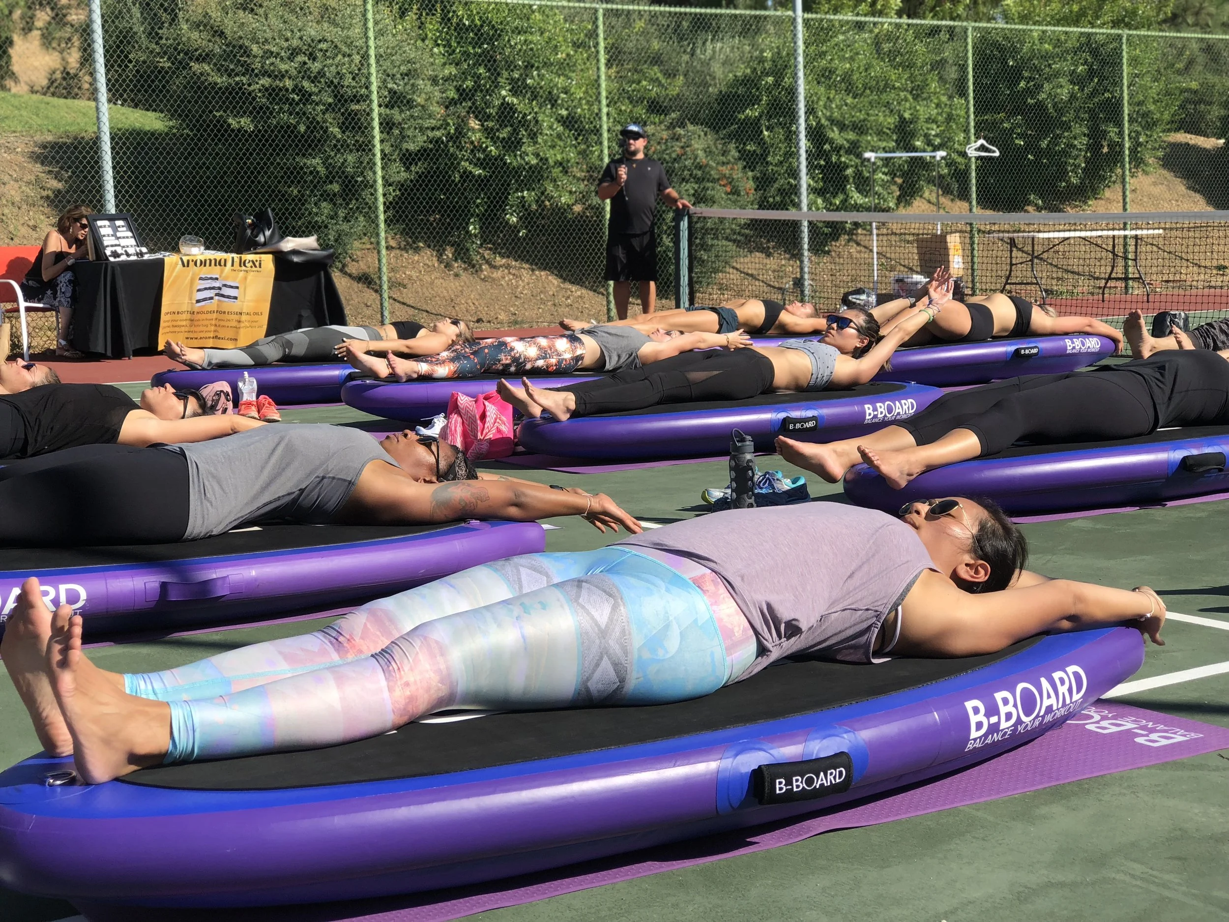 People participating in an outdoor b-board yoga class on purple balance boarads, lying on their backs with arms extended, in a group on a tennis court with a fence and trees in the background.