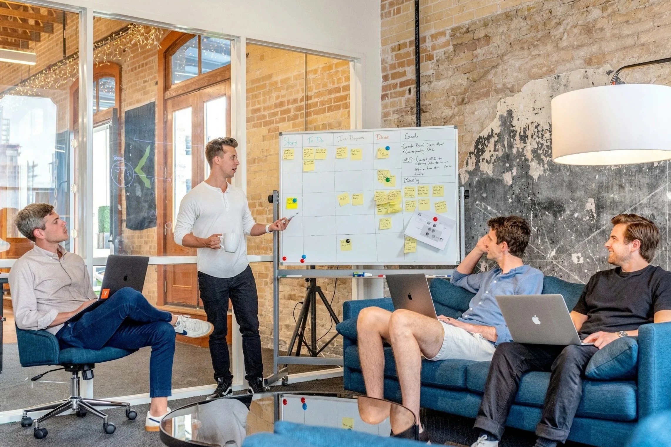 A group of people in a modern office or meeting room. One man stands near a whiteboard with sticky notes, holding a mug and pointing to the board. Others are seated on a sofa and chairs, listening. Windows with brick framing and string lights are visible.