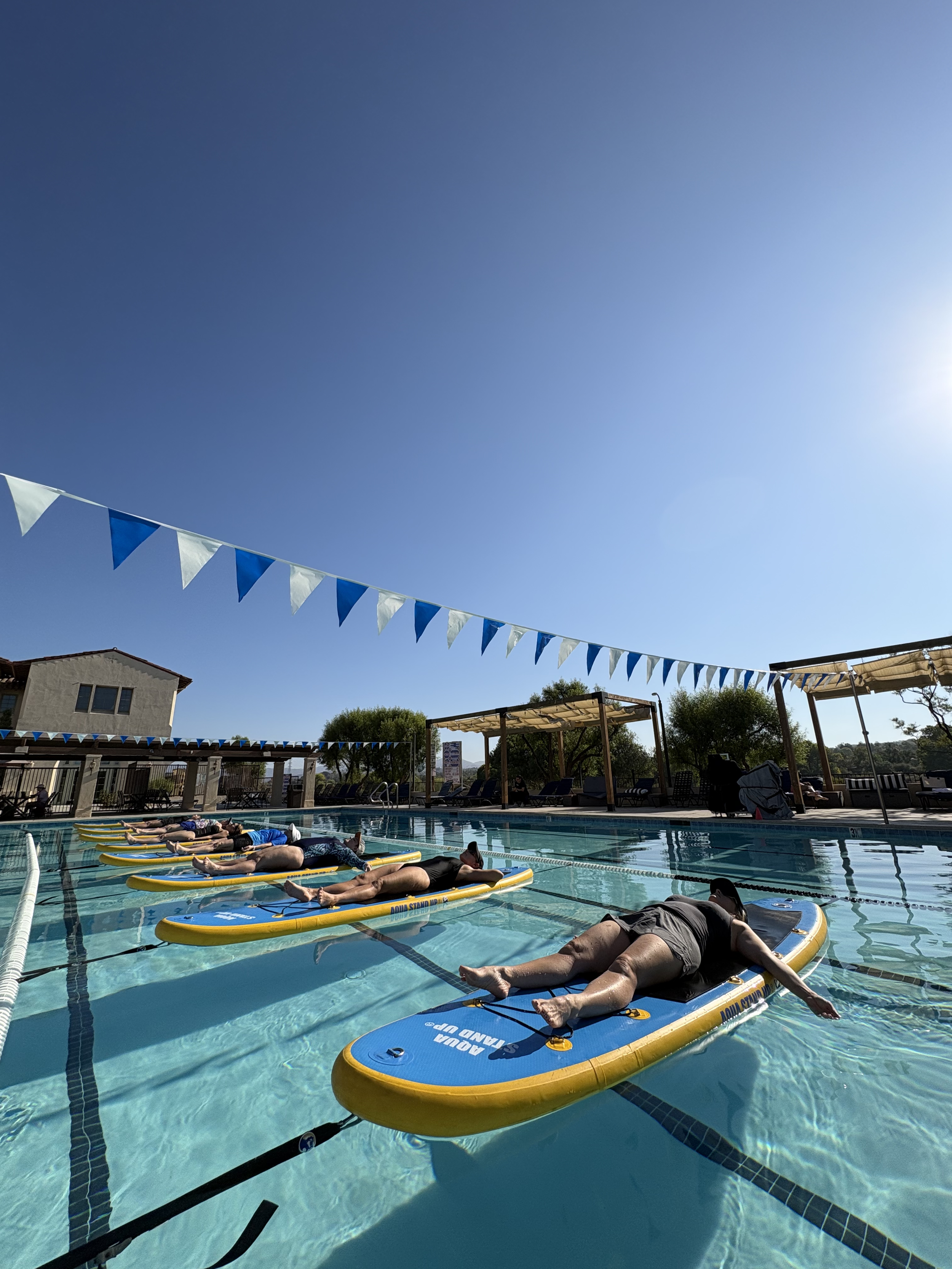 People relaxing on paddleboards in a swimming pool with blue and white triangular flags overhead on a sunny day.